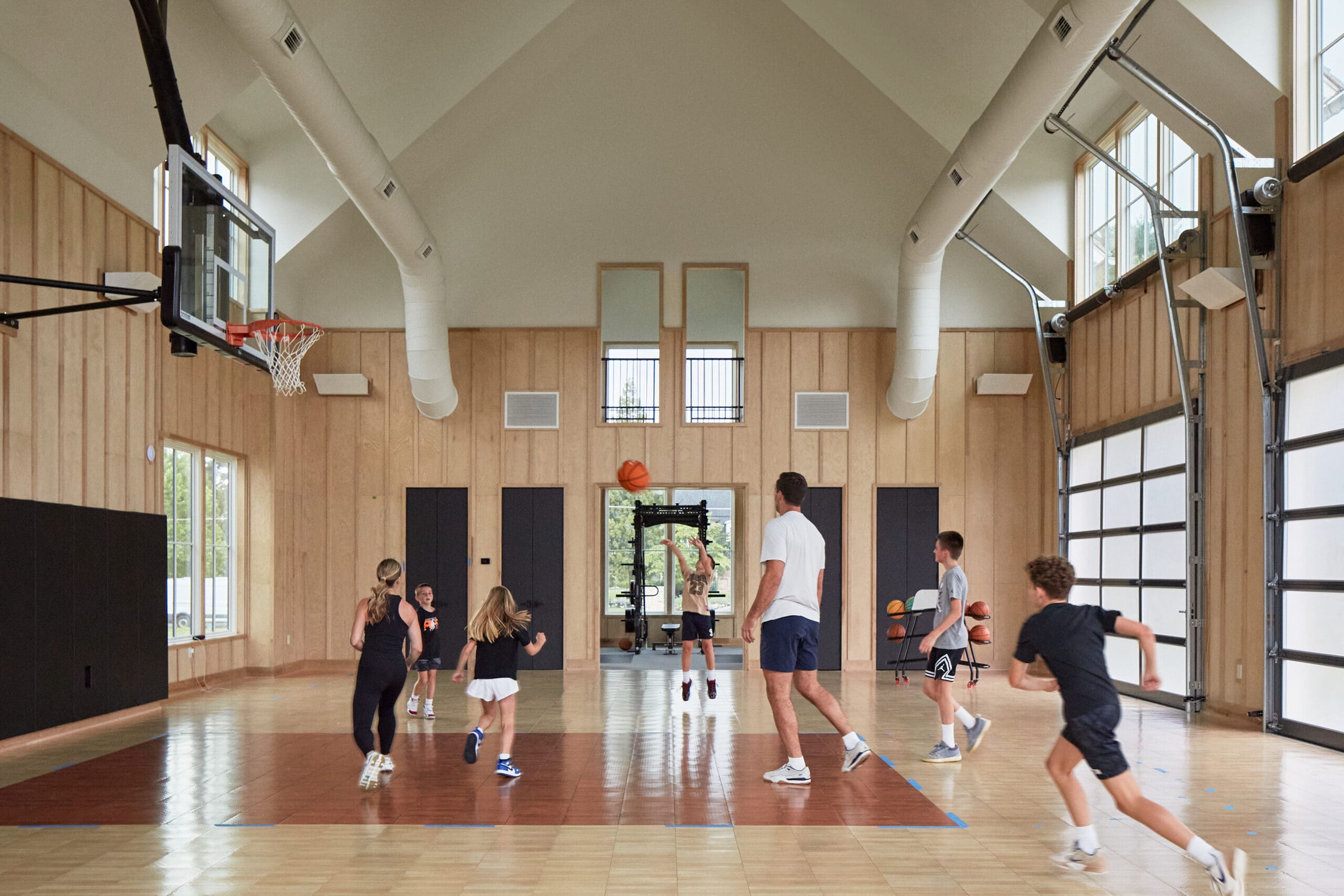 Indoor gym with players playing basketball near the hoop on the left, wooden walls and large windows in the background