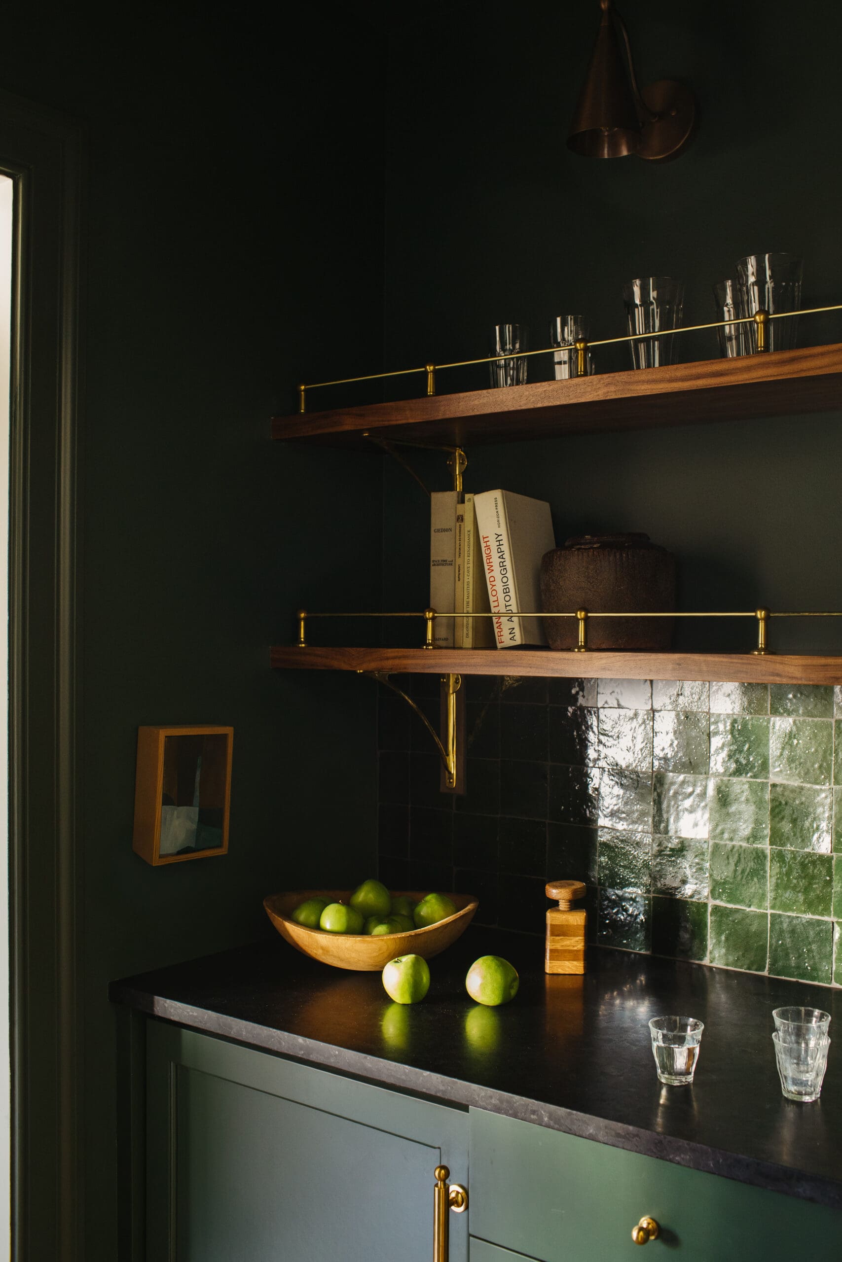 Moody kitchen scene with dark green walls, wooden shelves and brass rails, glass tumblers, books, a ceramic pot, and a bowl of limes on a black countertop.