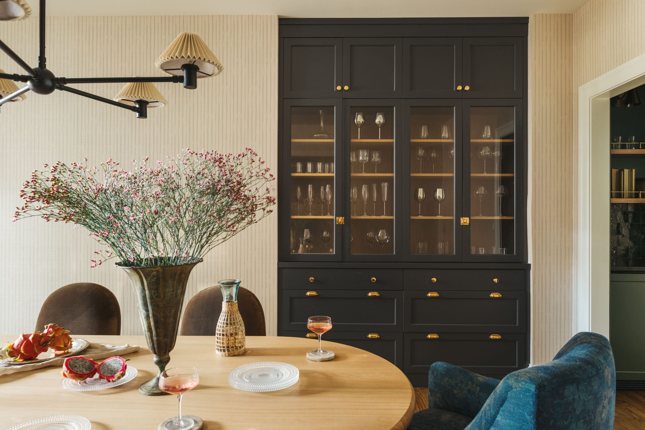 Dining room with a tall black glass-front cabinet, gold knobs, and a round wooden table set with glasses and a floral centerpiece.
