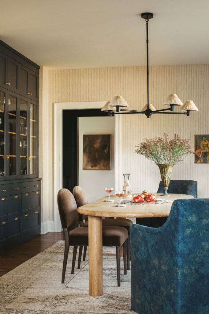 Elegant dining room with a black cabinet, striped wallpaper, and a round wooden table set with glasses and flowers under a black chandelier with beige lampshades.