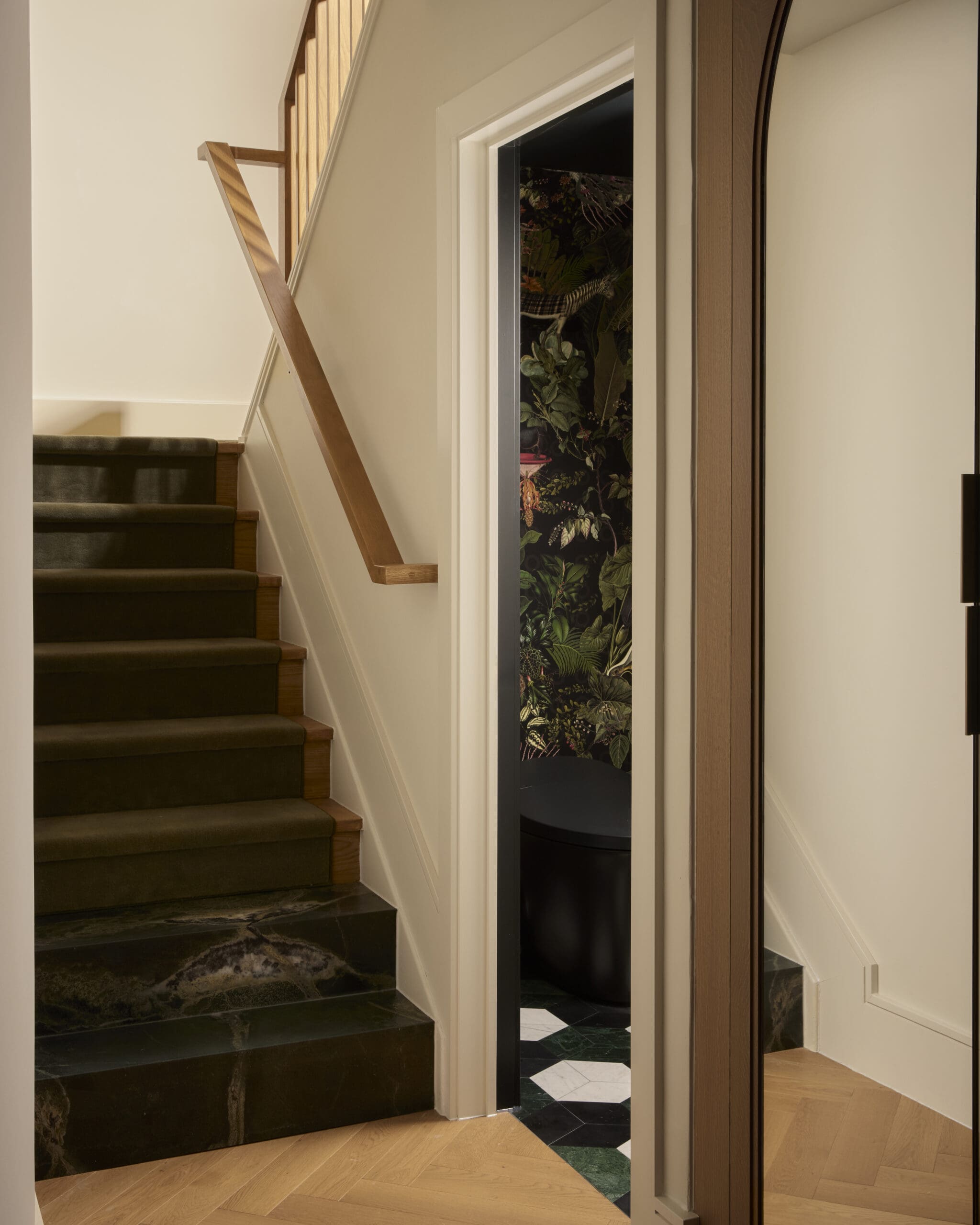 Wooden staircase with dark steps and light walls, railing on the left, leading up beside a doorway to a tropical‑leaf patterned room behind a gray door frame.