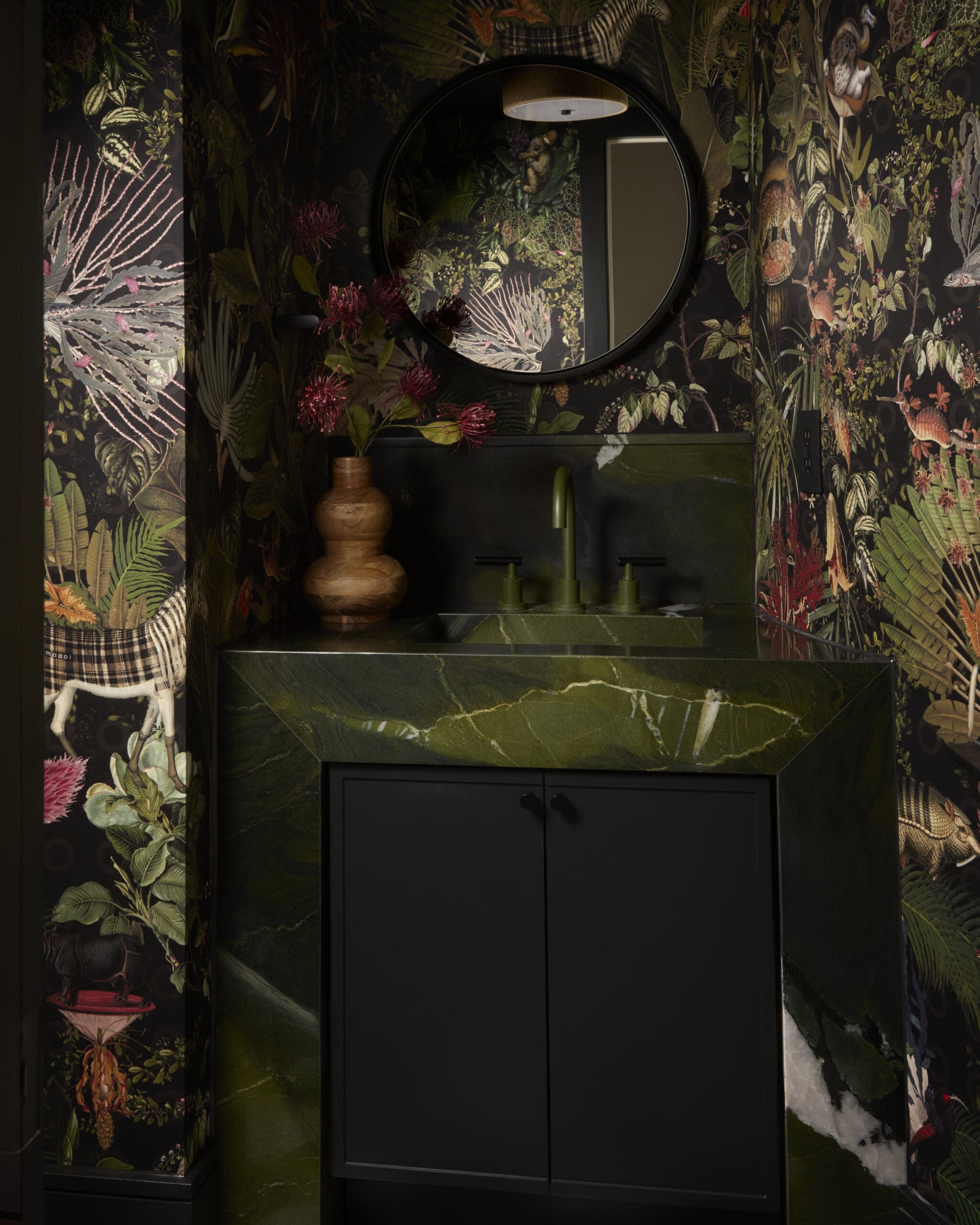 Dark tropical bathroom vanity with a round mirror, black cabinet, and green marble countertop against botanical wallpaper.