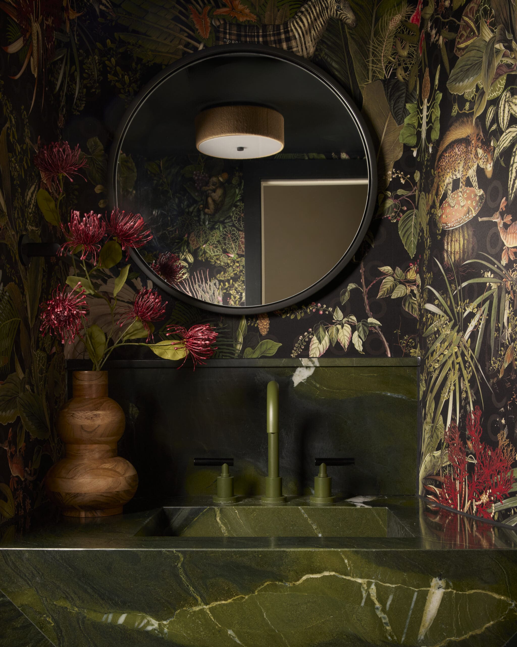 Round mirror on a tropical botanical wallpaper wall, reflecting a wooden ceiling light and dark green stone counter.