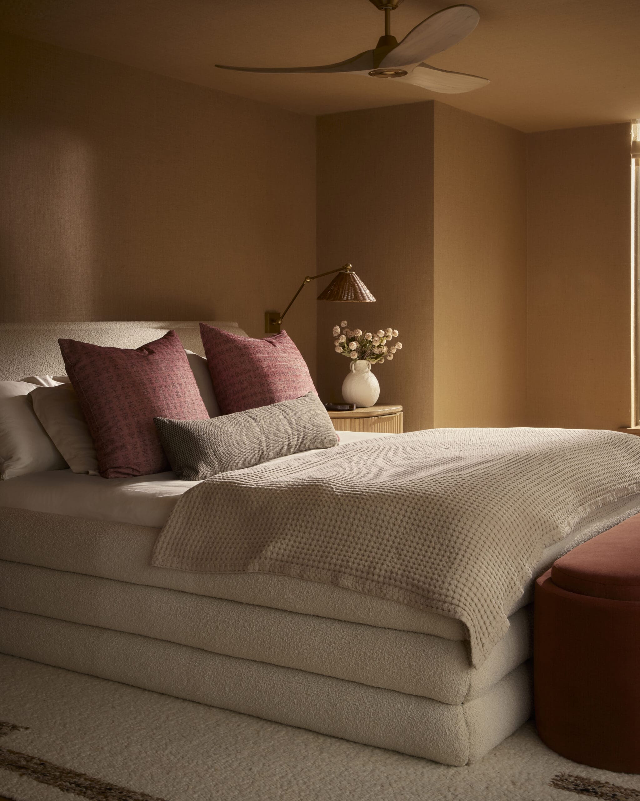 Neutral-toned bedroom with a plush bed, beige textured comforter, and pink patterned throw pillows; a round wooden nightstand holds a brass lamp and a vase of white flowers; ceiling fan overhead.