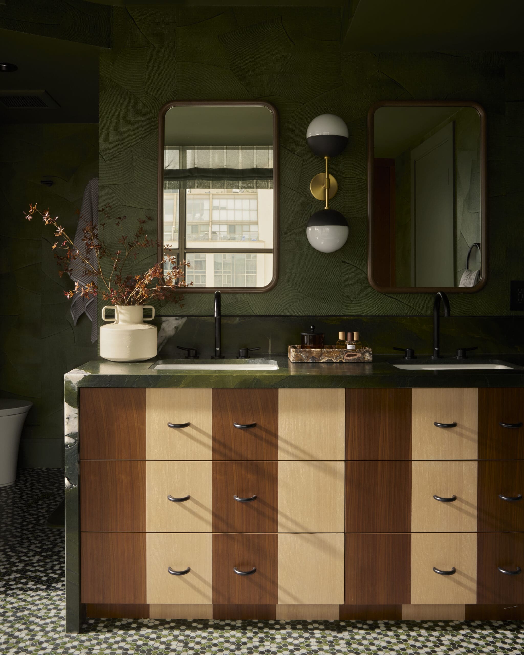 Dark green textured wall with two brown-framed mirrors, a gold-and-black globe wall light, and a marble-topped vanity with a soap dispenser and vase of branches.