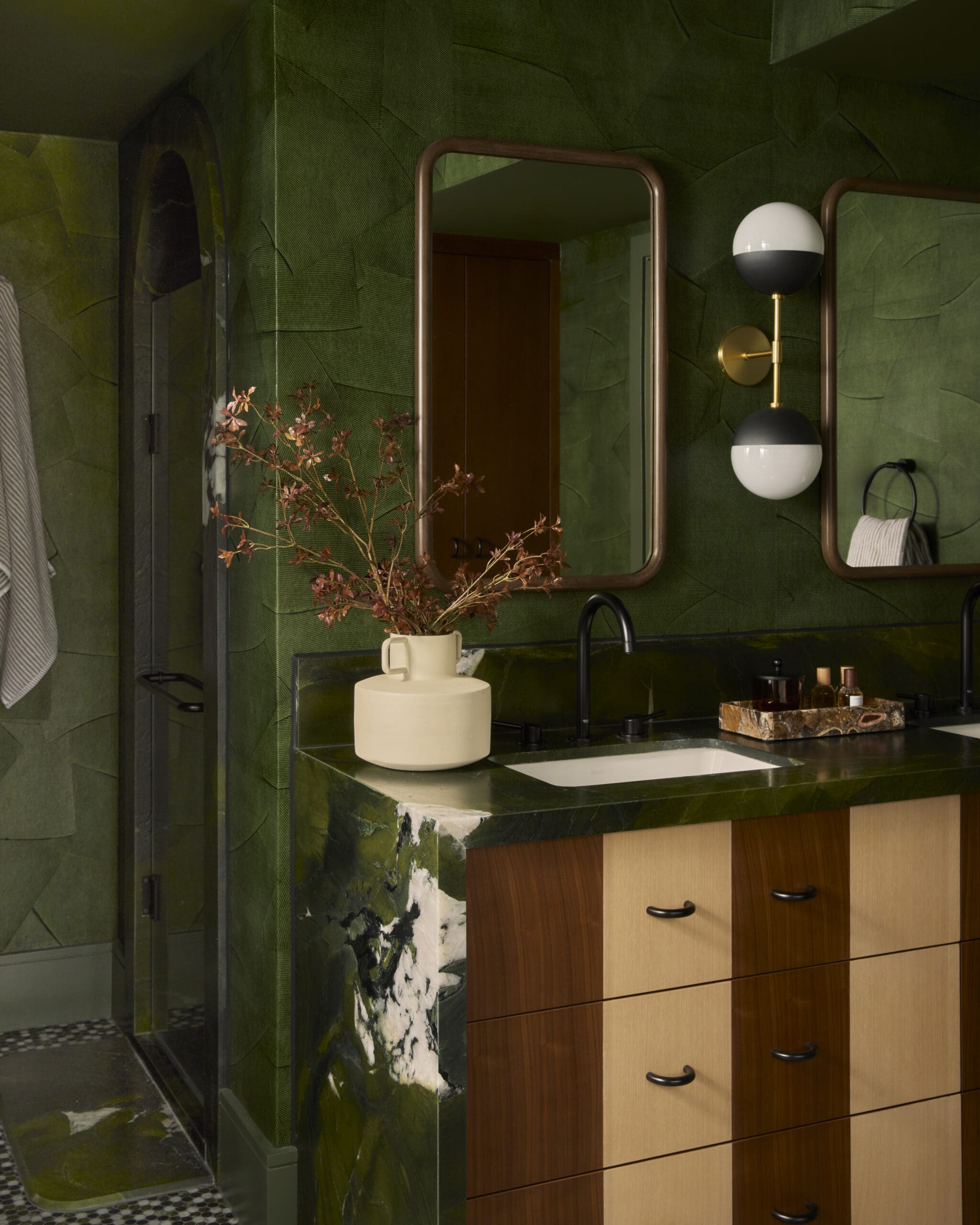 Emerald green bathroom with wooden vanity drawers, marble countertop, and round-edged mirrors reflecting a wooden door.