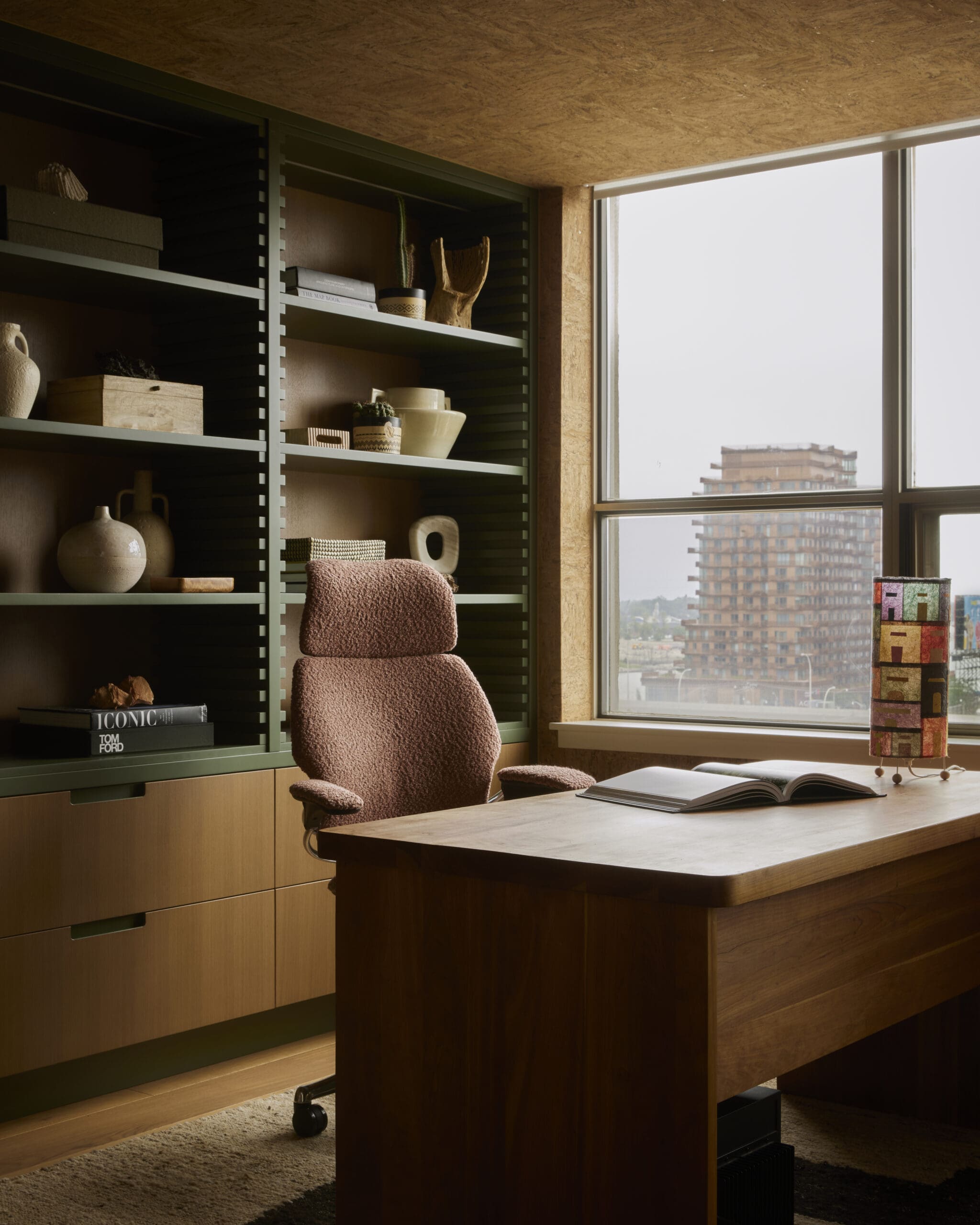 Office workspace with a wooden desk and burgundy chair beside built-in green shelves filled with pottery and books by a large window.