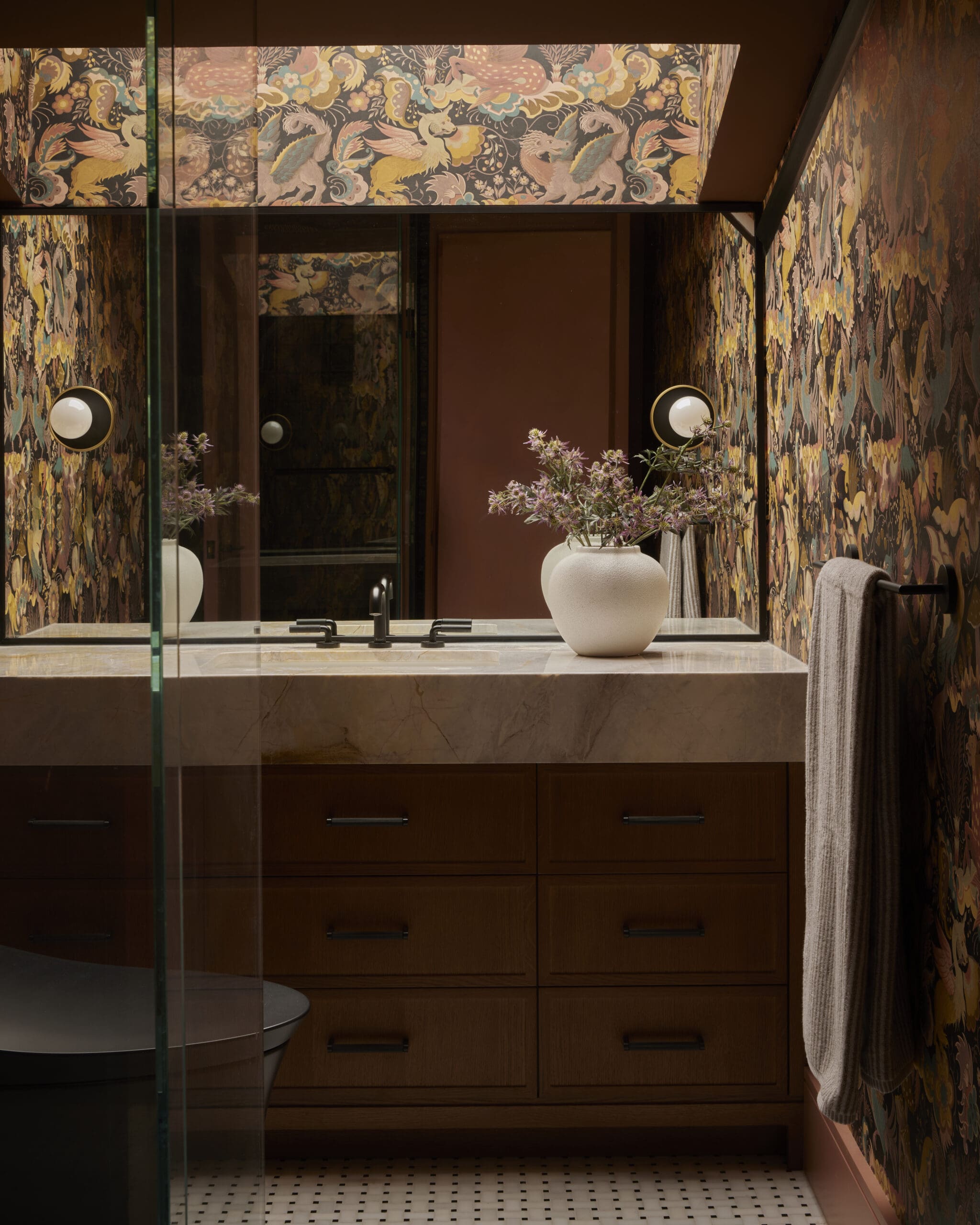 Bathroom vanity with dark wood drawers, marble countertop, and a white vase of purple flowers, set against colorful patterned wallpaper.