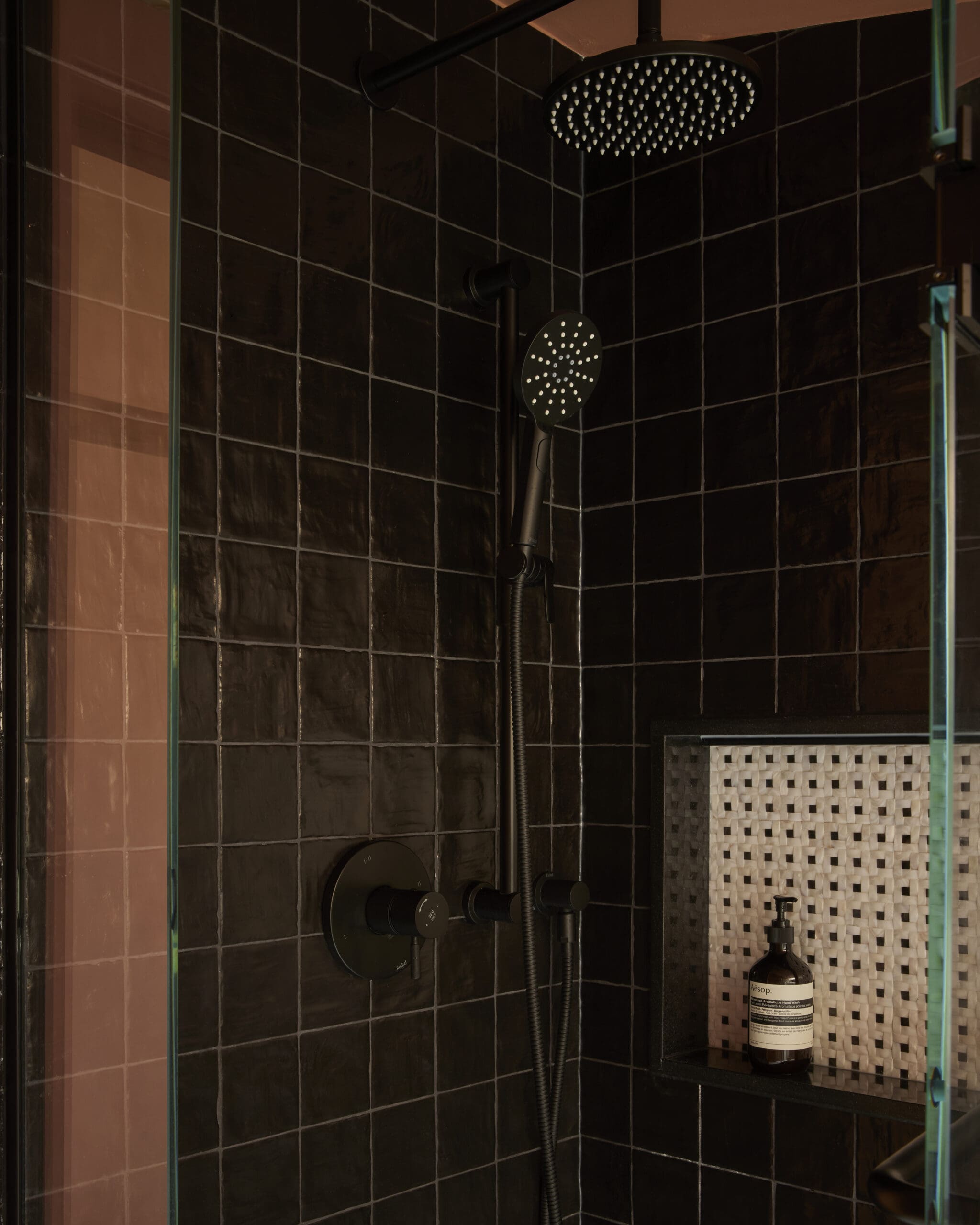 Shower area with dark tiled walls, a large round rainfall showerhead and a handheld spray on a rail, glass shower door, and a bottle on a tiled shelf.