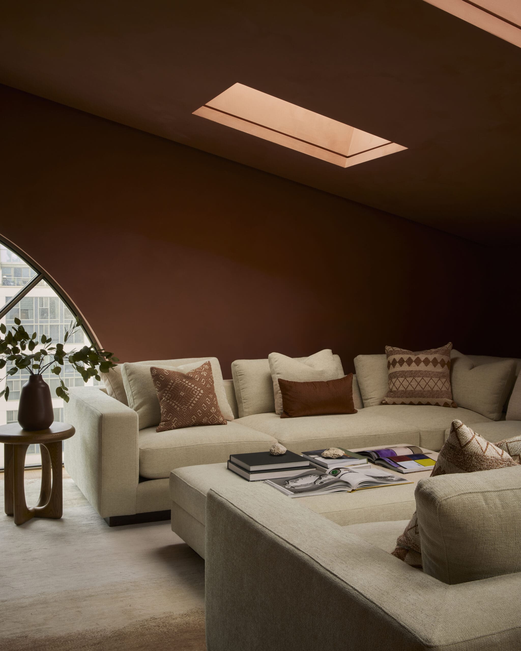 Beige sectional sofa with patterned pillows in a warm living room; magazines on the coffee table and a round vase on the side table near a curved window.