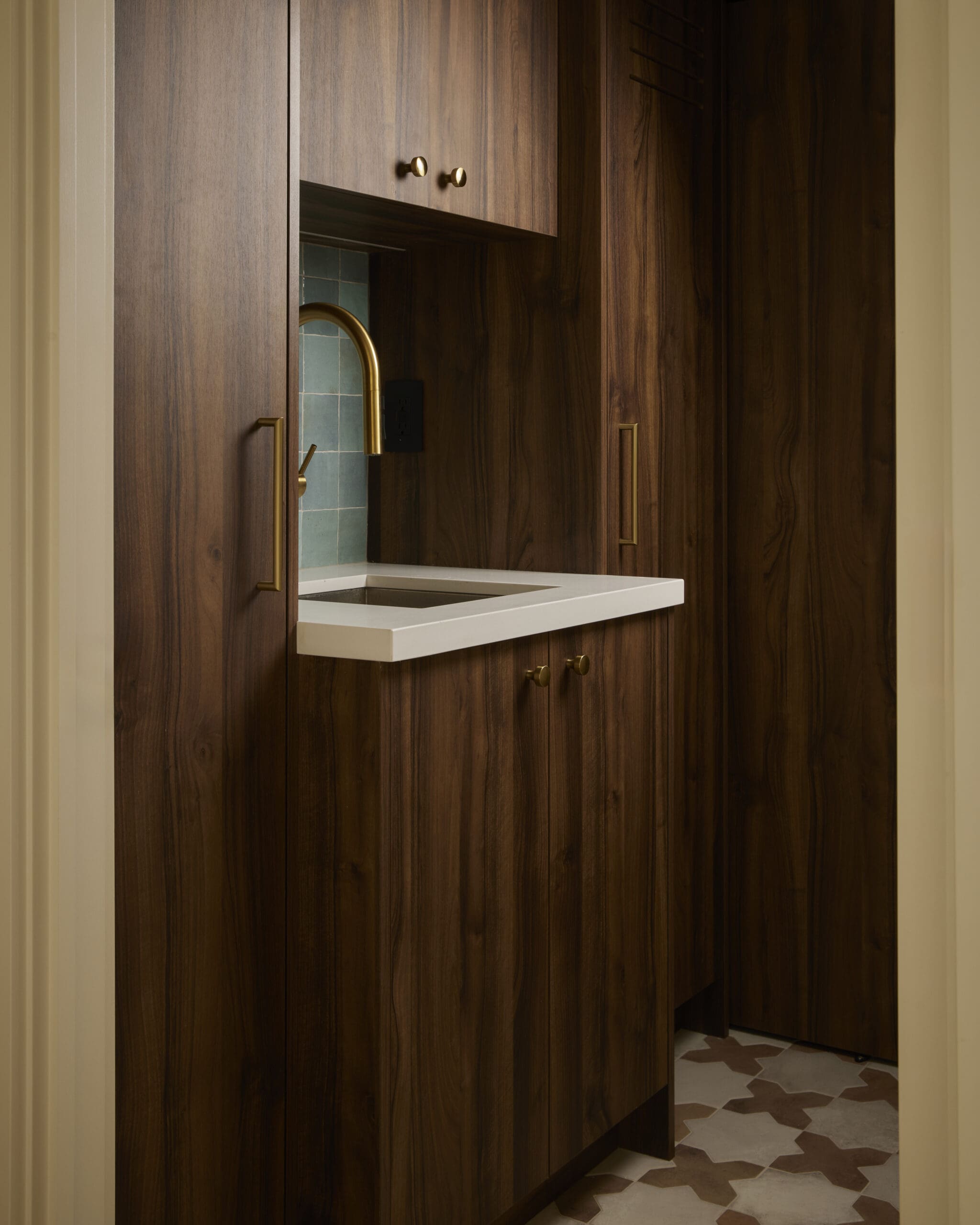 Brown wood kitchen cabinets with brass handles surrounding a teal-tiled backsplash and a gold faucet over a white counter/sink area.