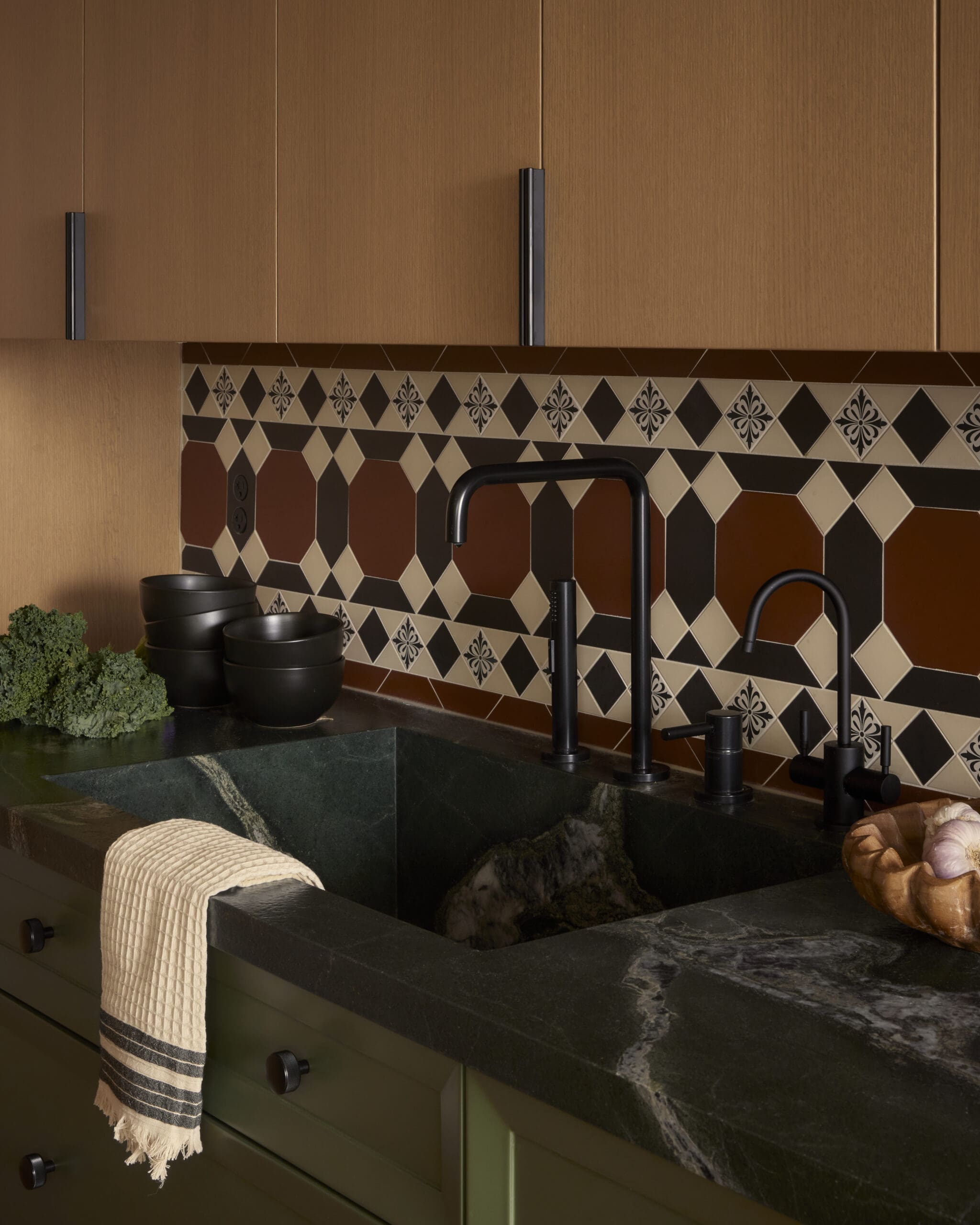 Black kitchen faucet over a dark stone countertop with a geometric tiled backsplash and wooden cabinets above.