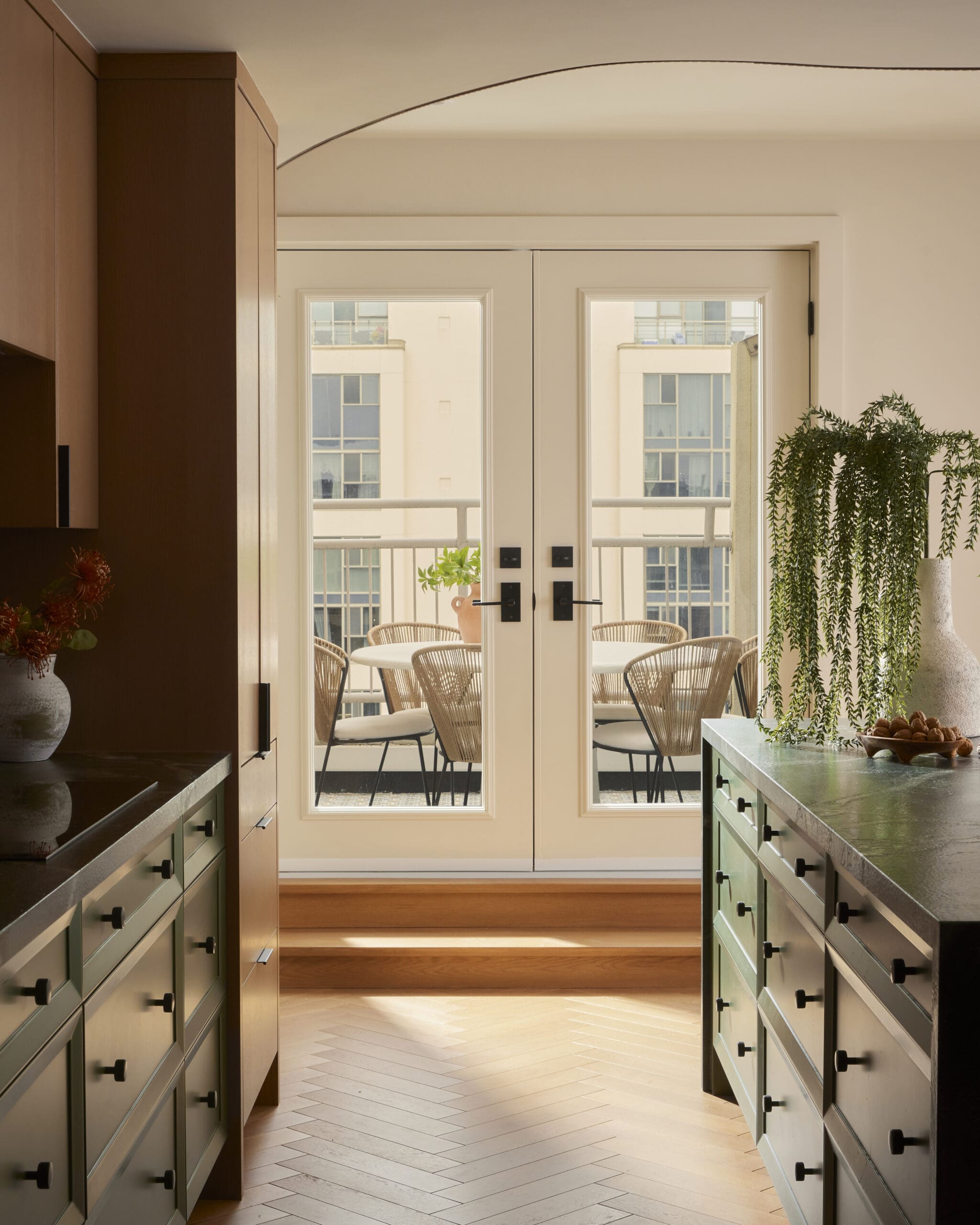 Kitchen corridor with brown cabinets on the left and mint-green drawers on both sides, opening to a balcony with wicker chairs outside.