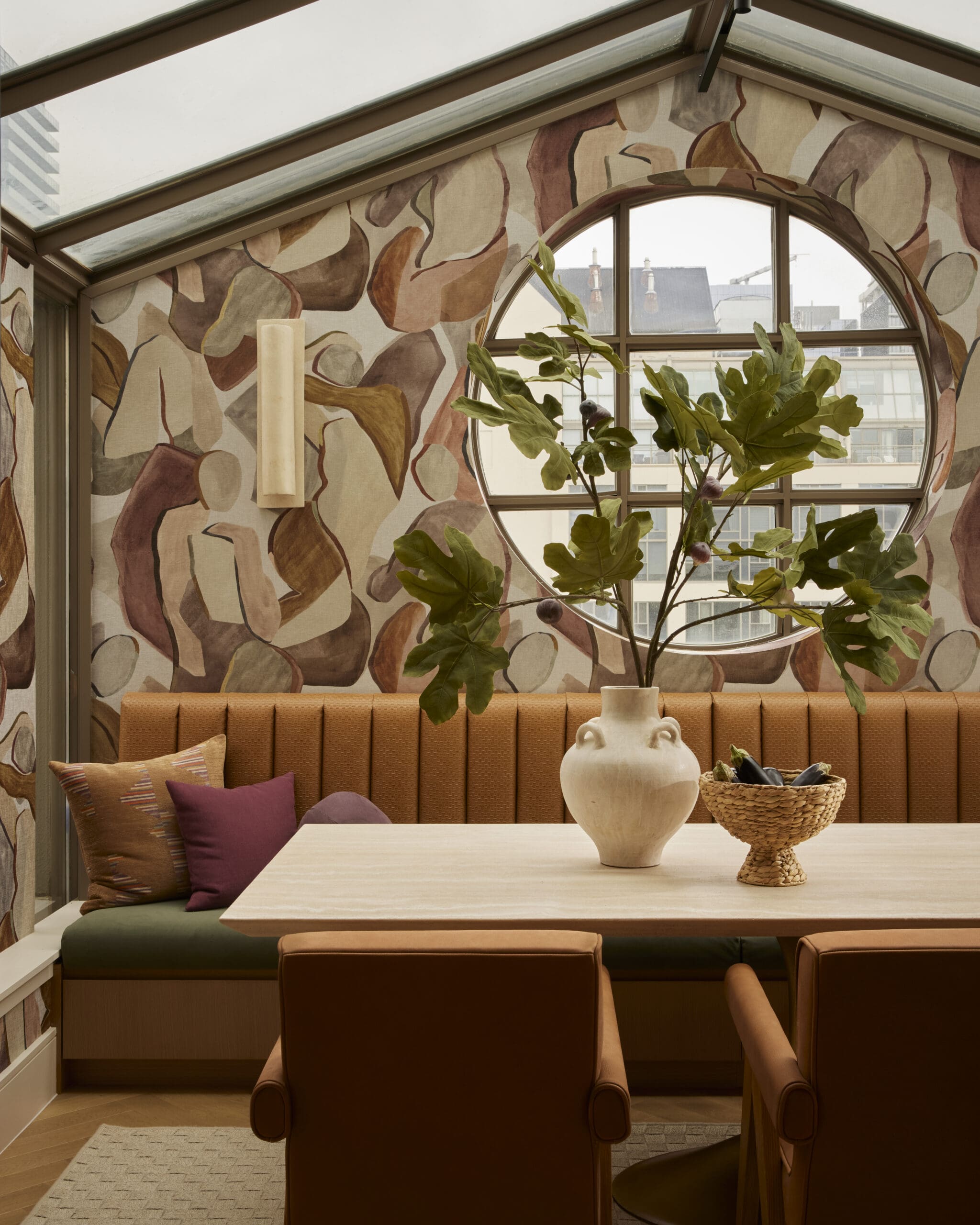 Cozy seating nook with orange-brown cushions, abstract patterned wallpaper, and a round window behind a leafy plant.