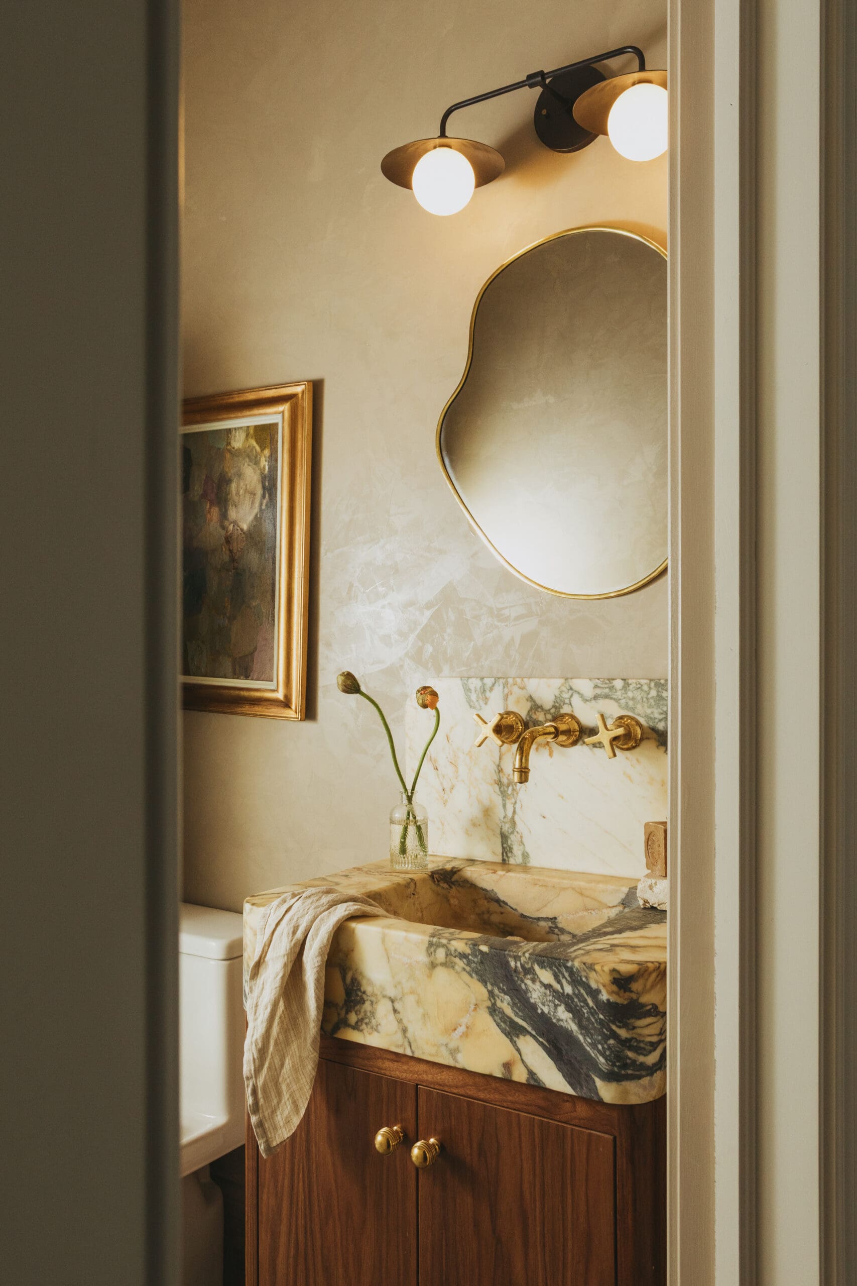 Elegant bathroom vanity with a marble countertop, brass hardware, and a round gold-framed mirror illuminated by warm wall sconces.