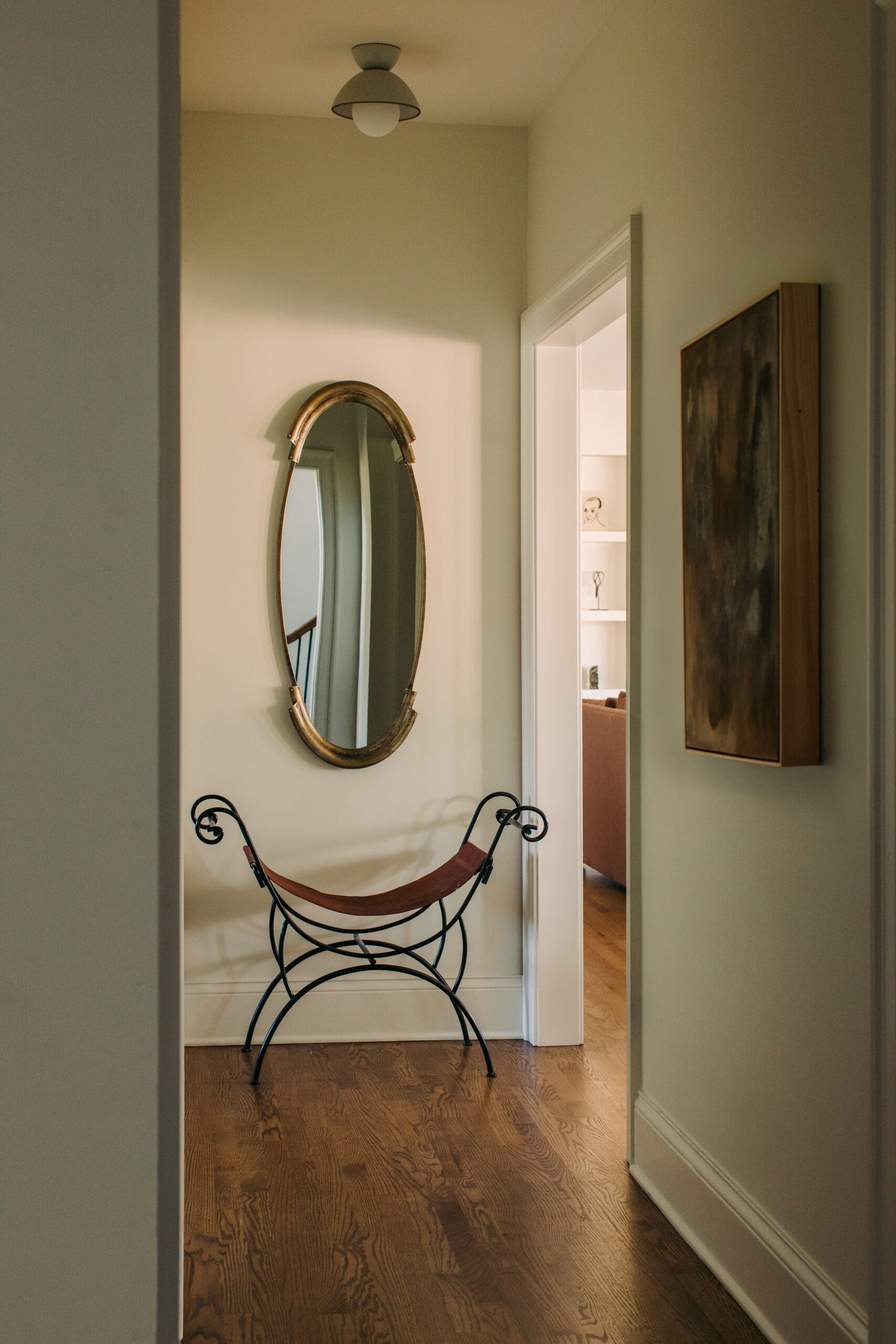 Narrow hallway nook with an oval gold mirror on the wall and a curved metal-framed hammock chair beneath it, warm lighting.