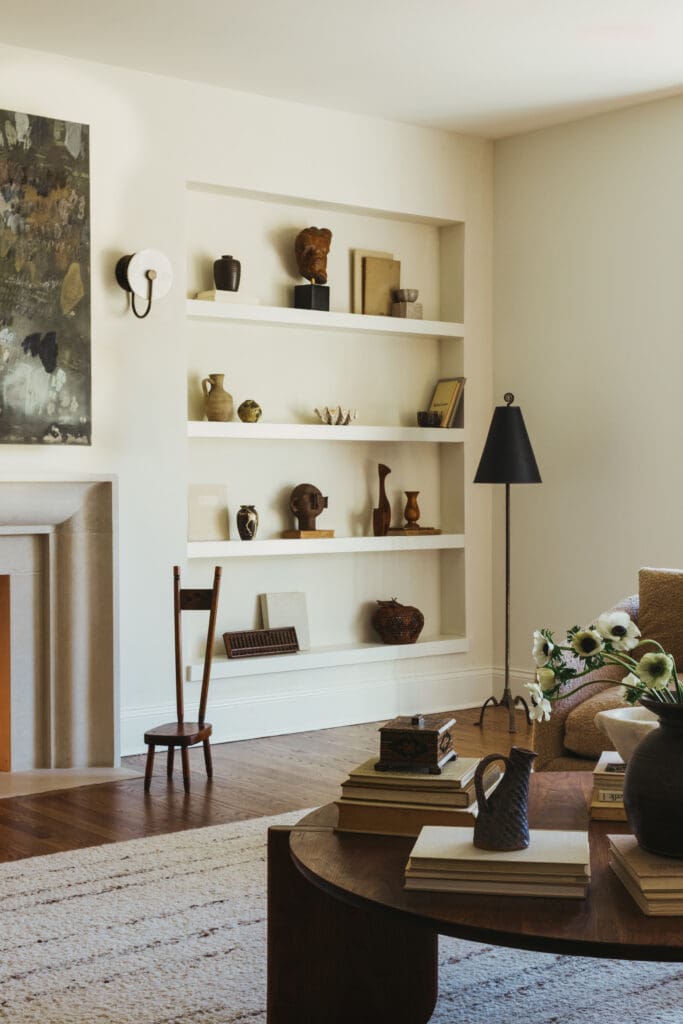 Living room with a white built-in shelf filled with pottery, books, and small sculptures, a black floor lamp nearby, and a round wooden coffee table in the foreground.