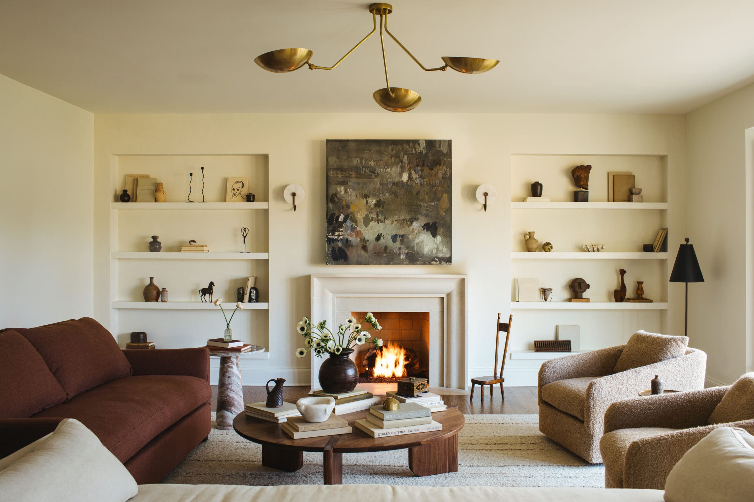 Cozy living room with built-in white shelves, a lit fireplace, and a brass chandelier above a central abstract painting.