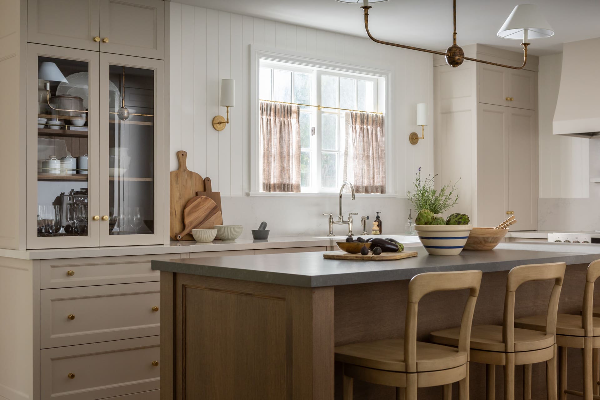 Cream-toned kitchen with a wooden island and brass hardware, bright window light at the sink area, and light curtains nearby.