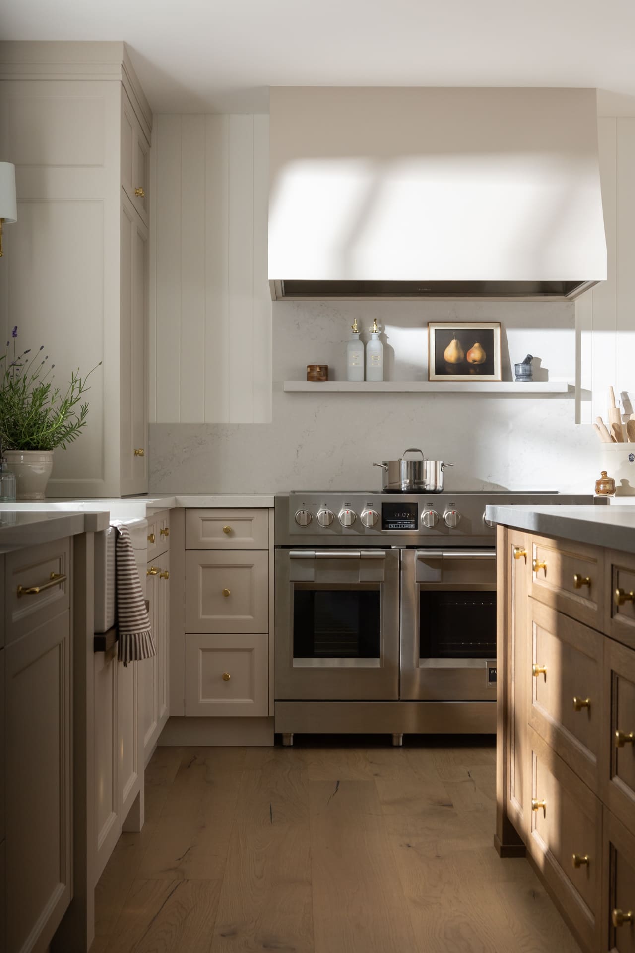 Cream kitchen with a stainless steel oven, marble backsplash, and a floating shelf displaying two bottles, a small copper container, and a pear still-life frame.