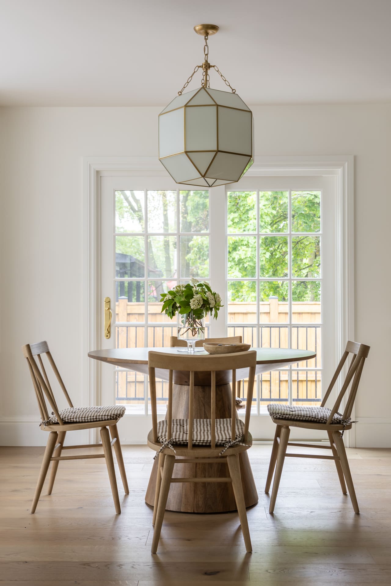 Round wooden dining table with four chairs and a vase of greenery in a bright dining nook, a geometric pendant light overhead and French doors to a sunny backyard