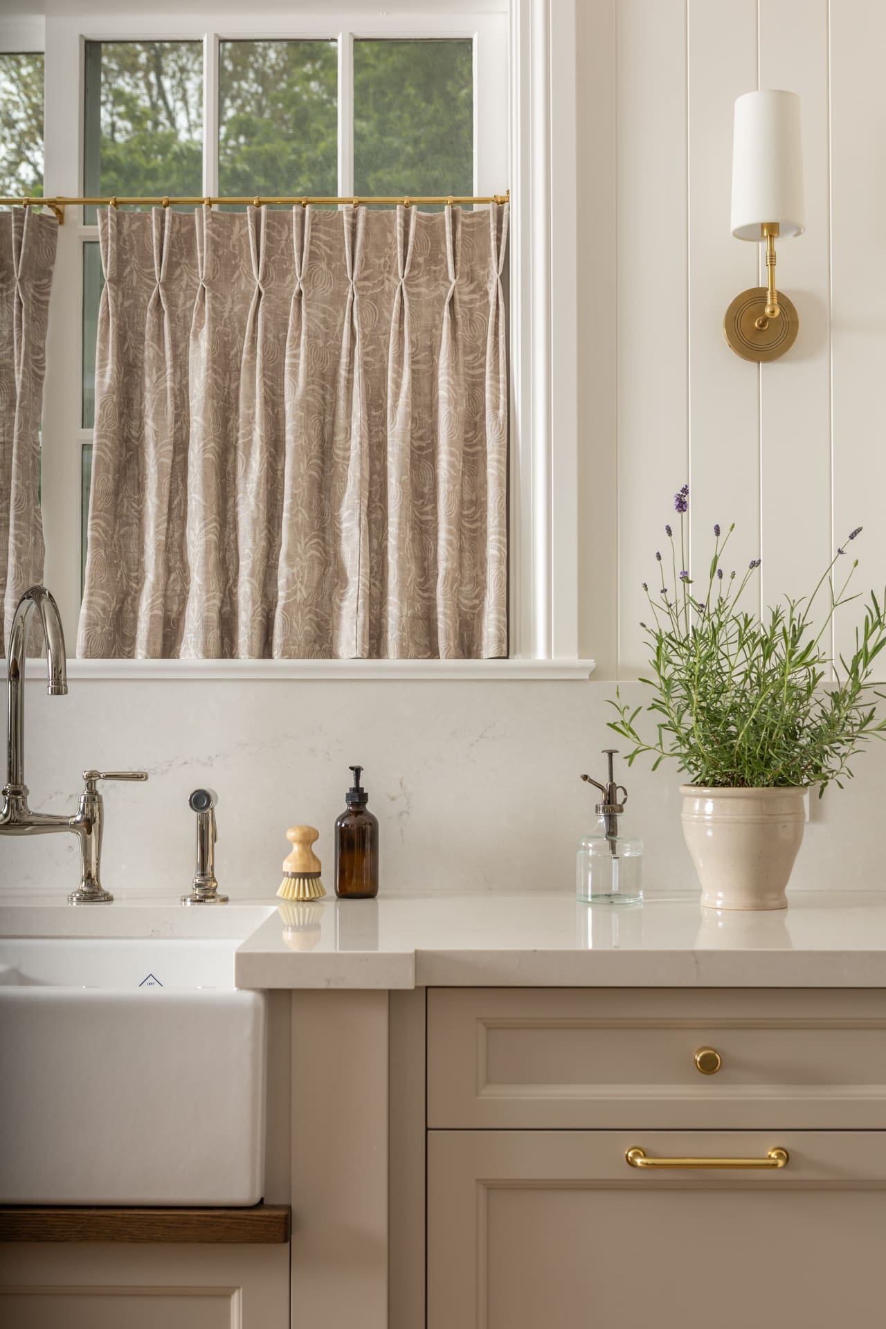 Kitchen counter with a white sink and brass faucet, beige cabinetry, and a potted lavender plant by a sunlit window with patterned beige curtains.