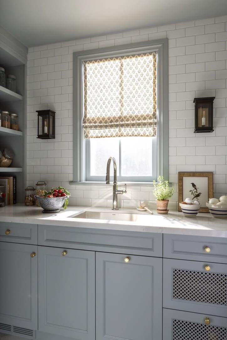 Kitchen sink area with light blue cabinets, white subway tile and a patterned window shade above the faucet.