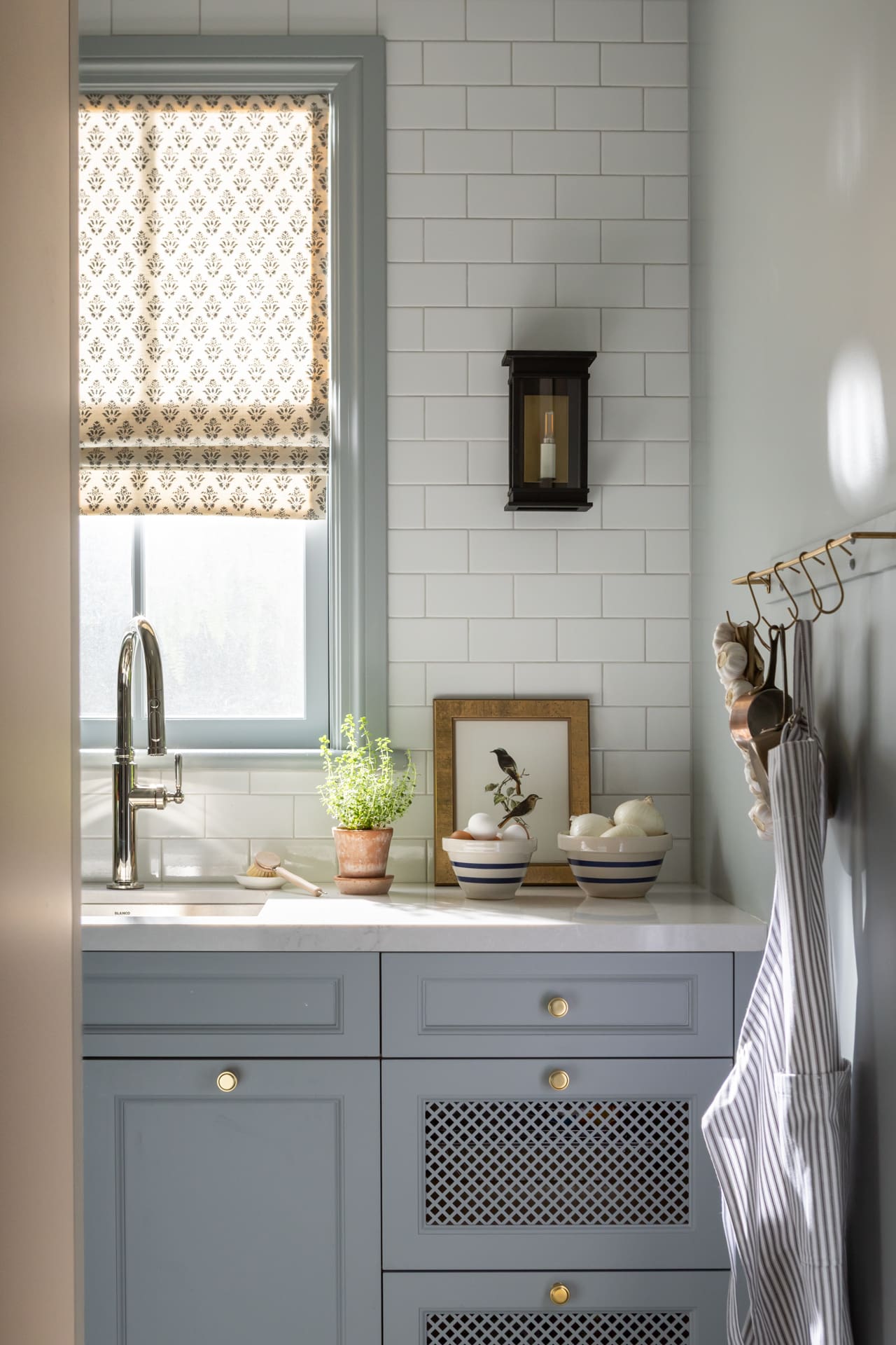 Blue-gray kitchen with brass hardware, white subway tile, a window with patterned shade, and a wall lantern.