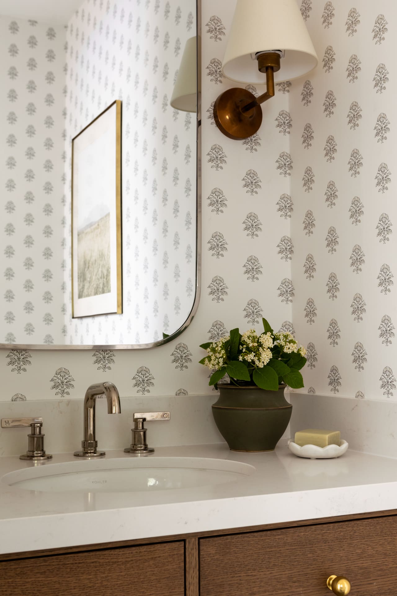 Bathroom vanity with marble countertop, dual chrome faucets, and a brass wall sconce above a mirror with patterned wallpaper; a vase of white flowers on the counter