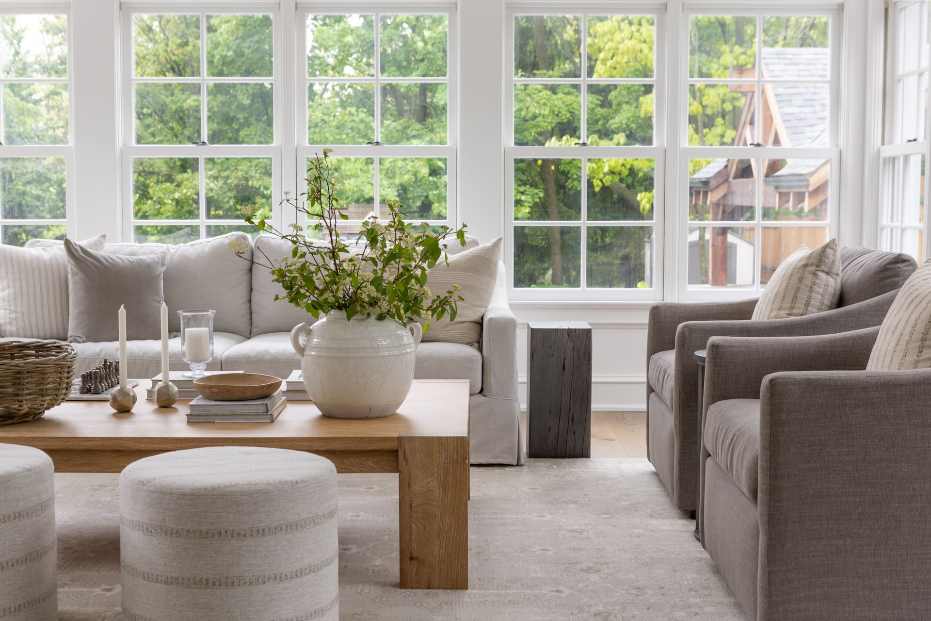 Bright living room with white sofa and gray chairs around a wooden coffee table, large multi-pane windows showing greenery outside.