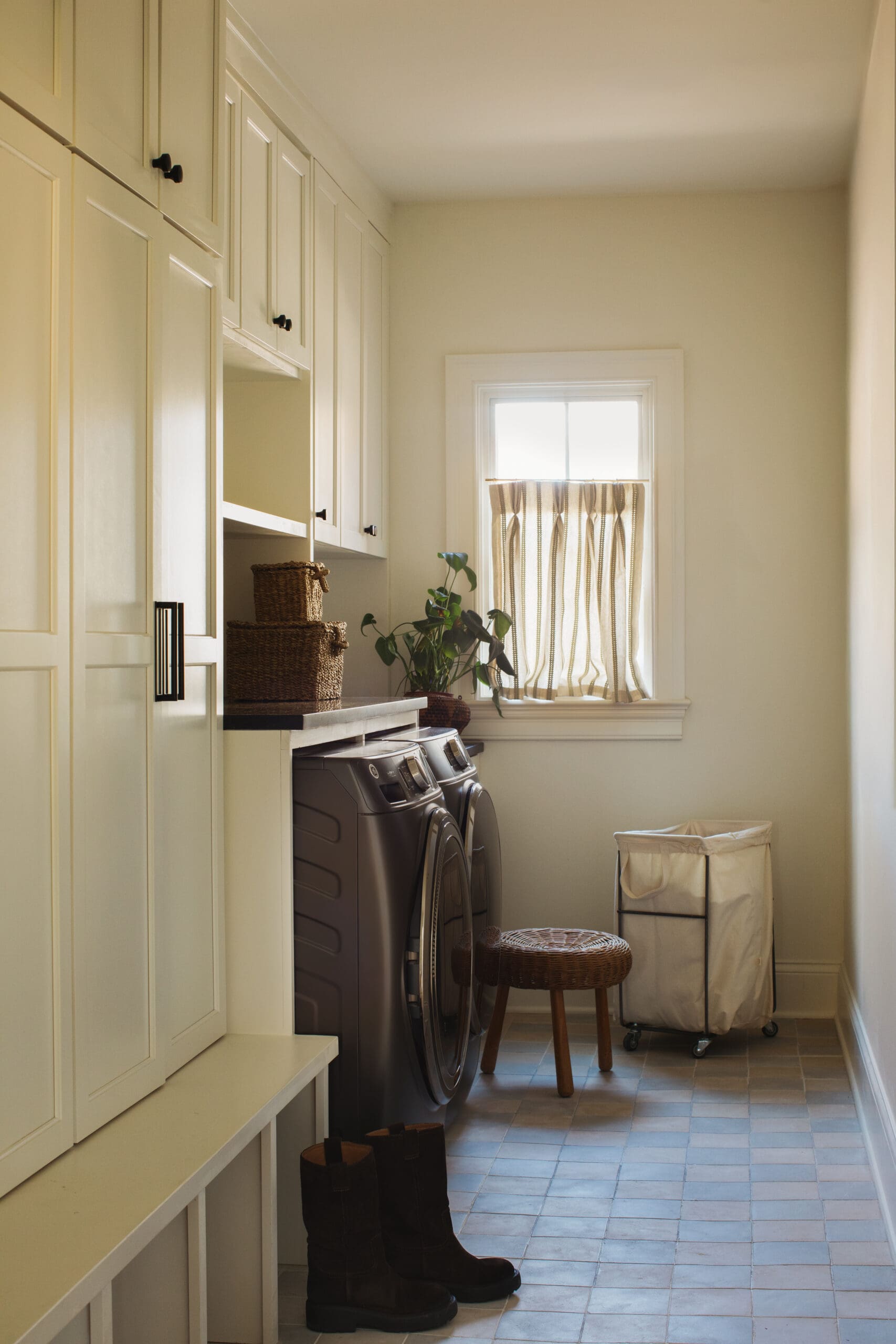 Laundry room with tall cream cabinets, front-loading washer and dryer, wicker baskets, a potted plant, and a window with striped curtain.