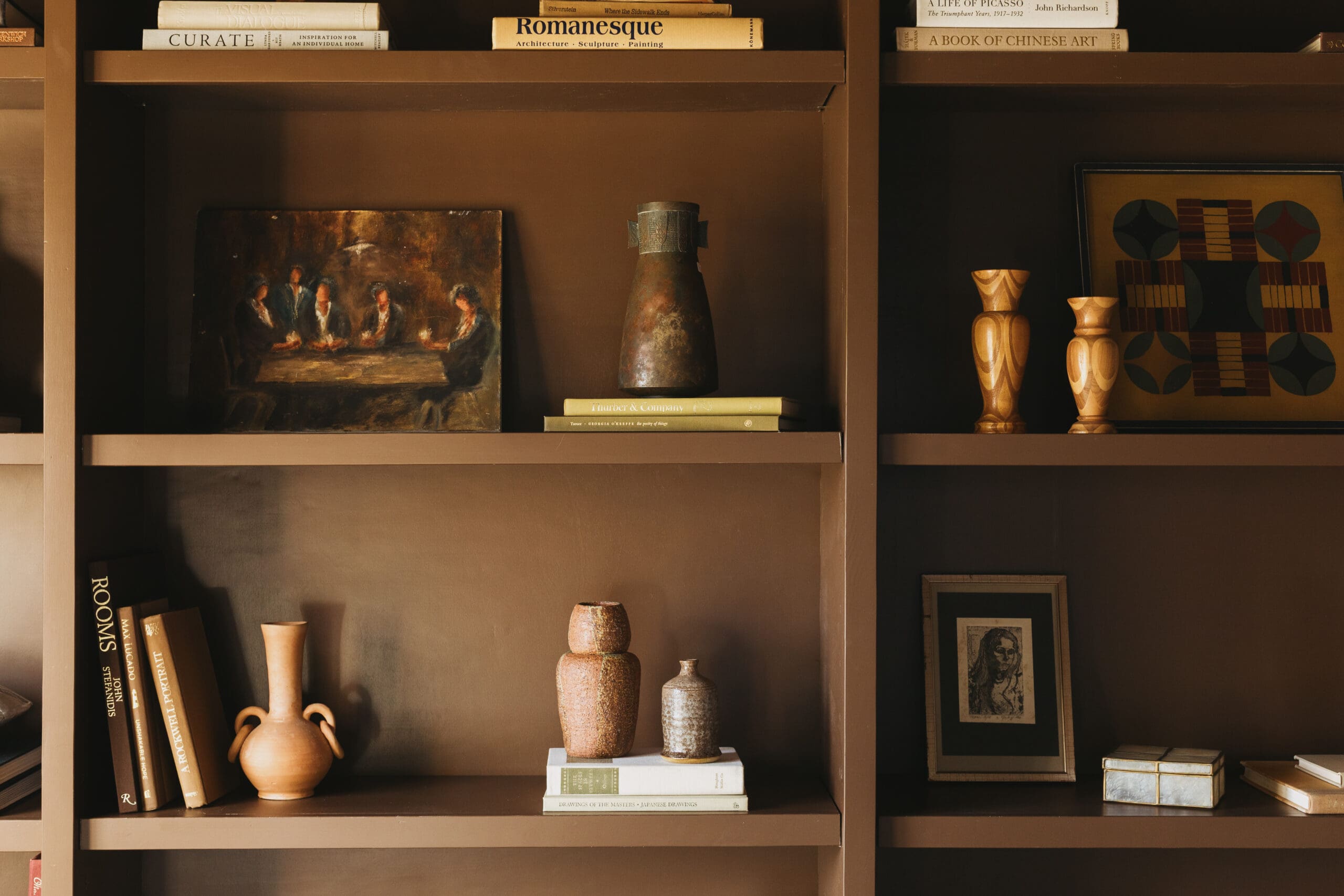 Brown shelving unit filled with books, ceramic vases, and framed art; middle shelf features a small oil painting of people at a table.