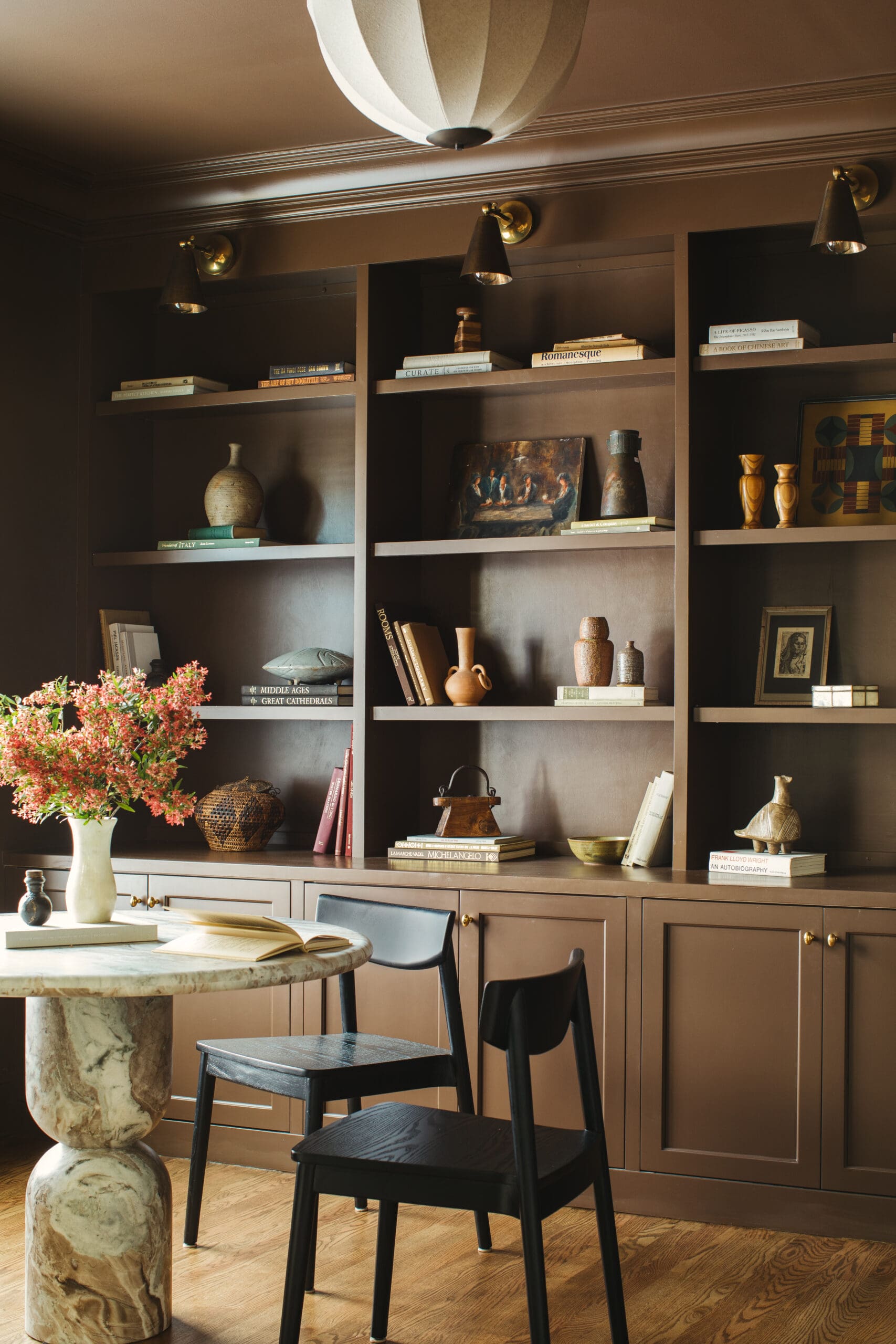 Brown built-in bookshelf filled with books and decorative objects, with a marble-topped round table and a vase of pink flowers in the foreground.