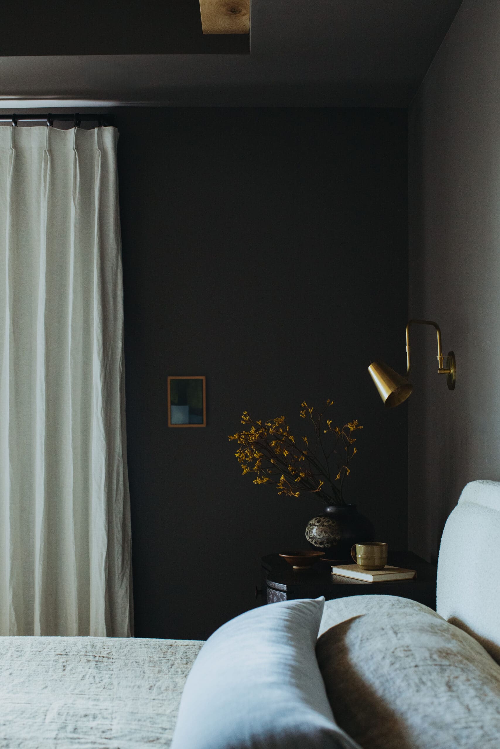 Dark living room corner with a brass wall lamp, cream curtains, and a round table holding a vase of dried branches beside a light sofa.