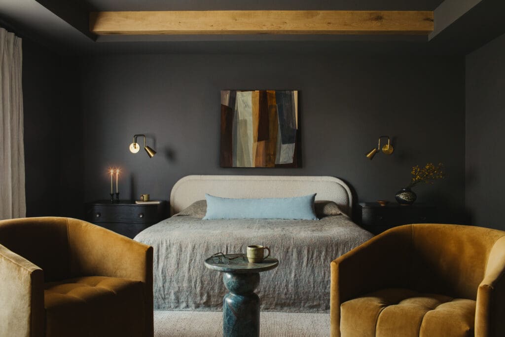Dark charcoal bedroom with upholstered bed, abstract painting above headboard, and two brass sconces flanking the bed.