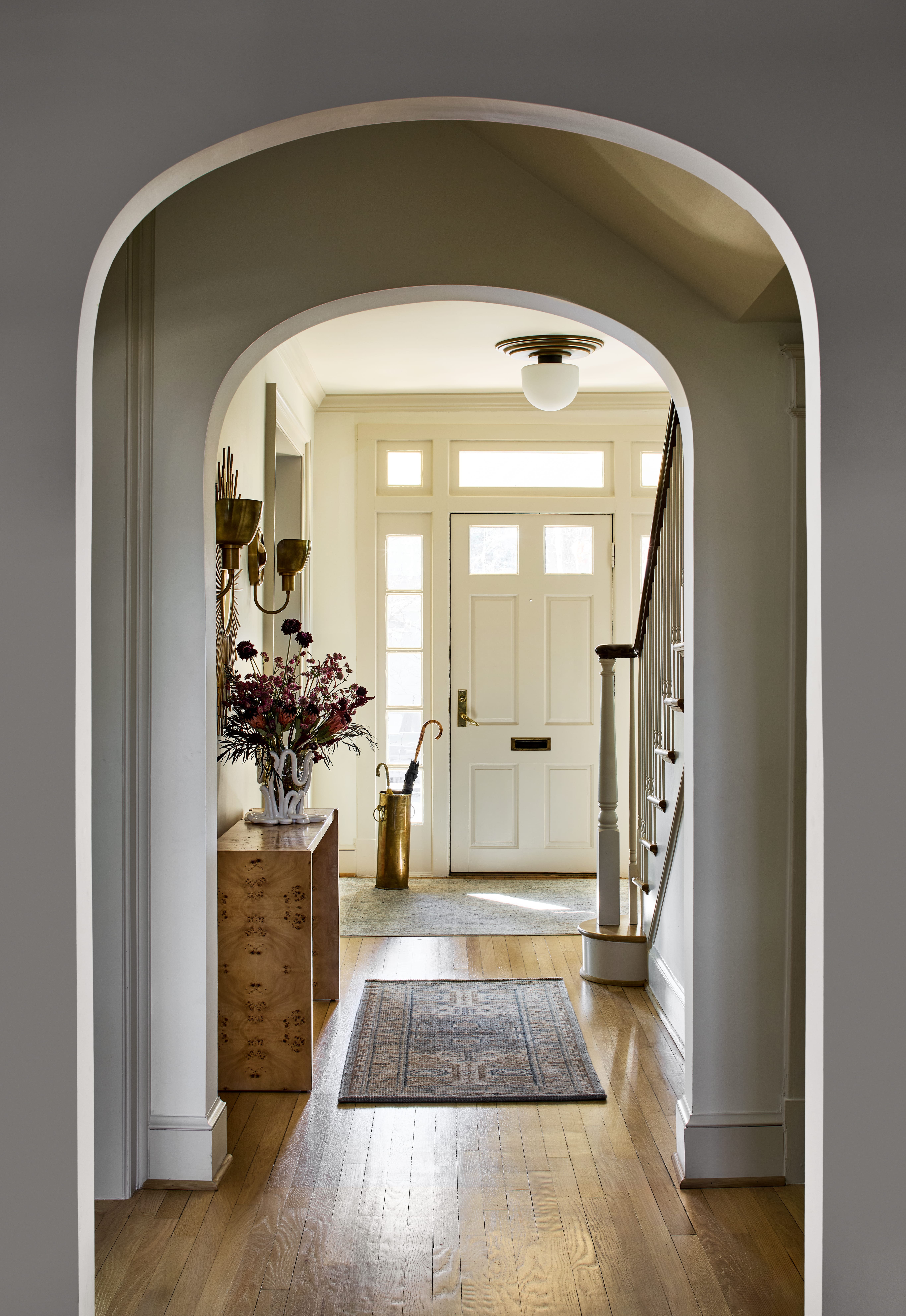 View down a warmly lit hallway framed by a rounded archway, front door at end, staircase to the right, and a wooden console with a vase of flowers.