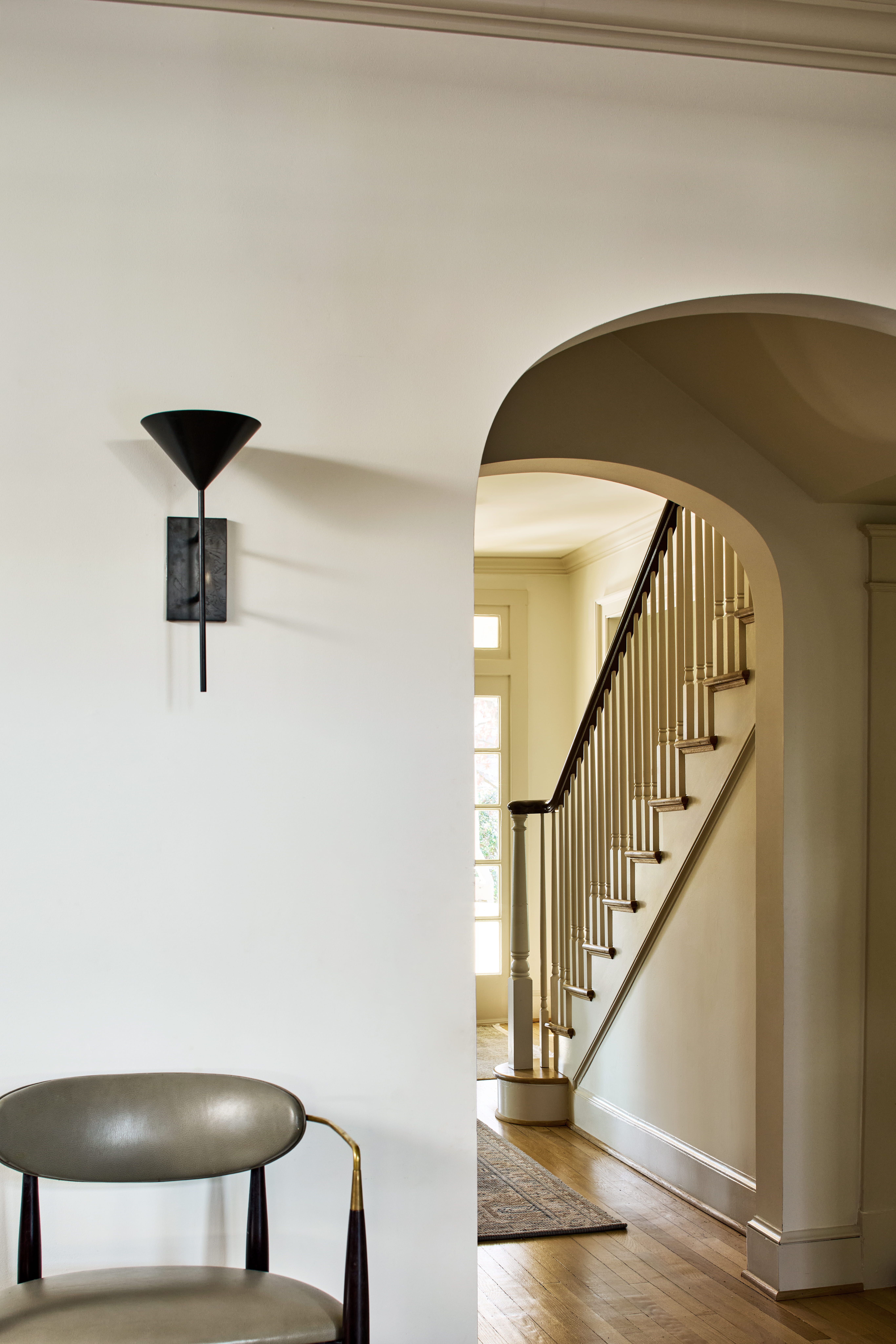 White wall with a black cone-shaped wall lamp and an arched doorway revealing a wooden staircase with white balusters on the right, plus a partial chair in the foreground.