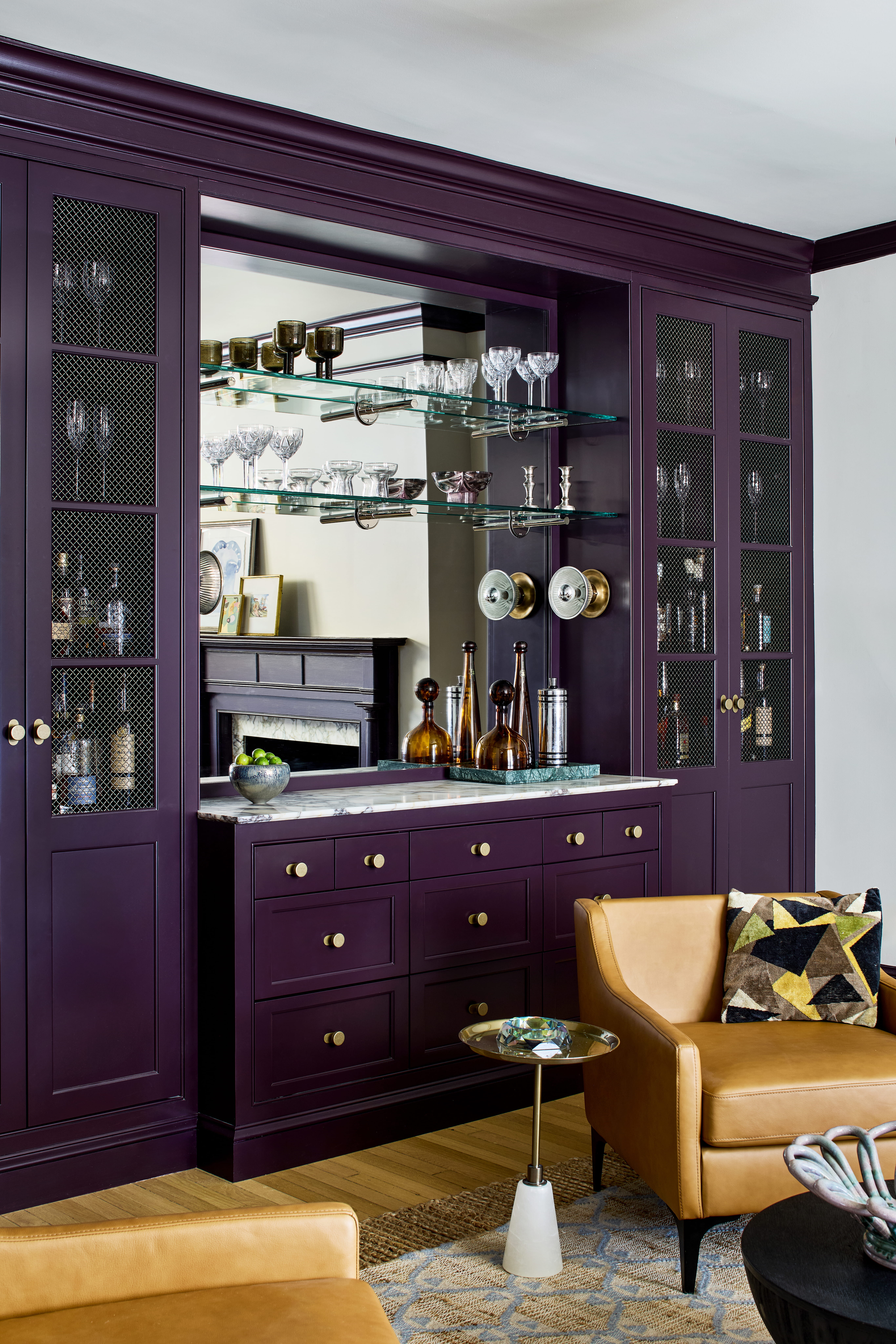 A luxurious home bar with purple cabinetry, glass shelves and crystal glassware arranged above a marble countertop.
