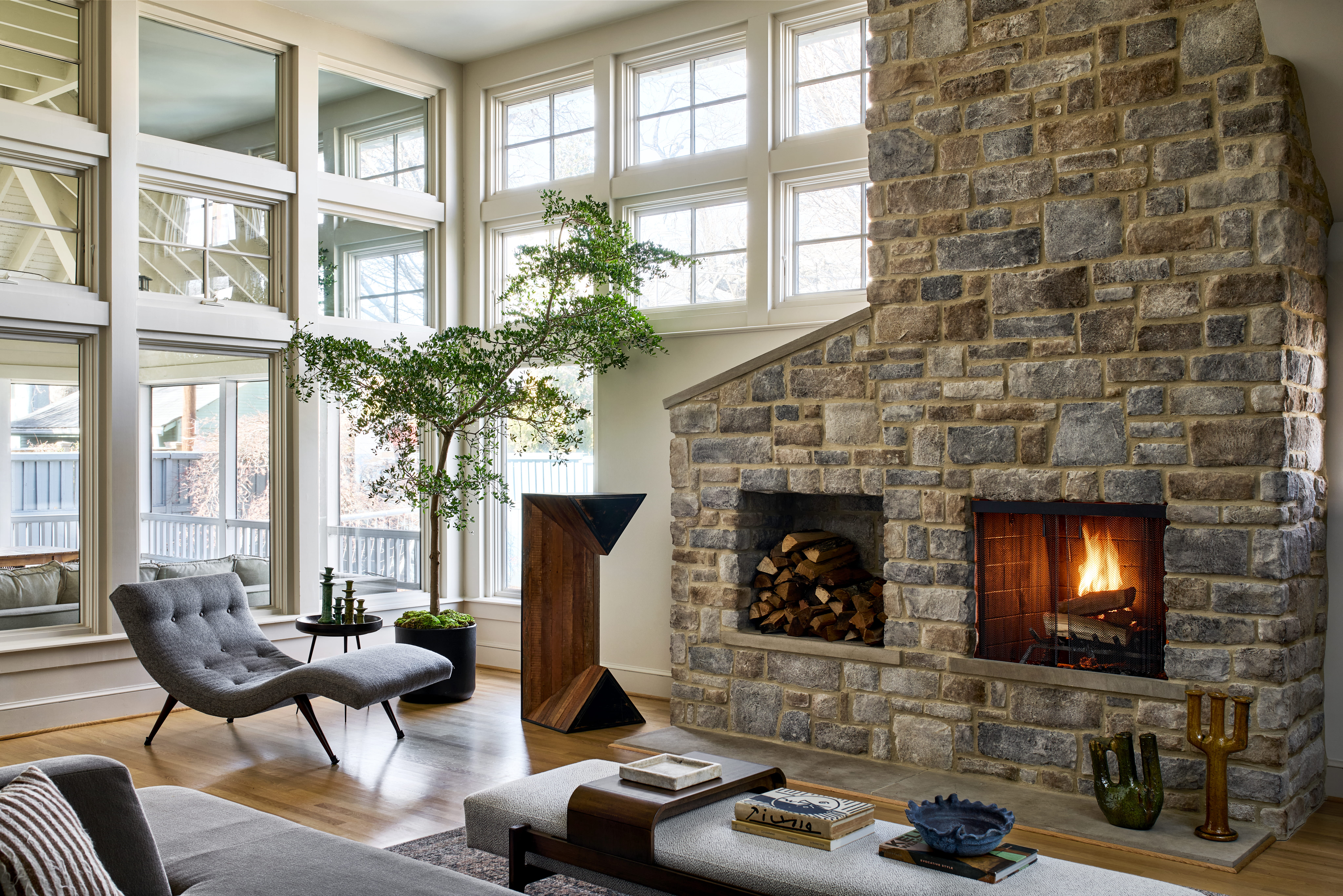 Cozy living room with a large stone fireplace, tall windows, and a green potted tree by a gray lounge chair.