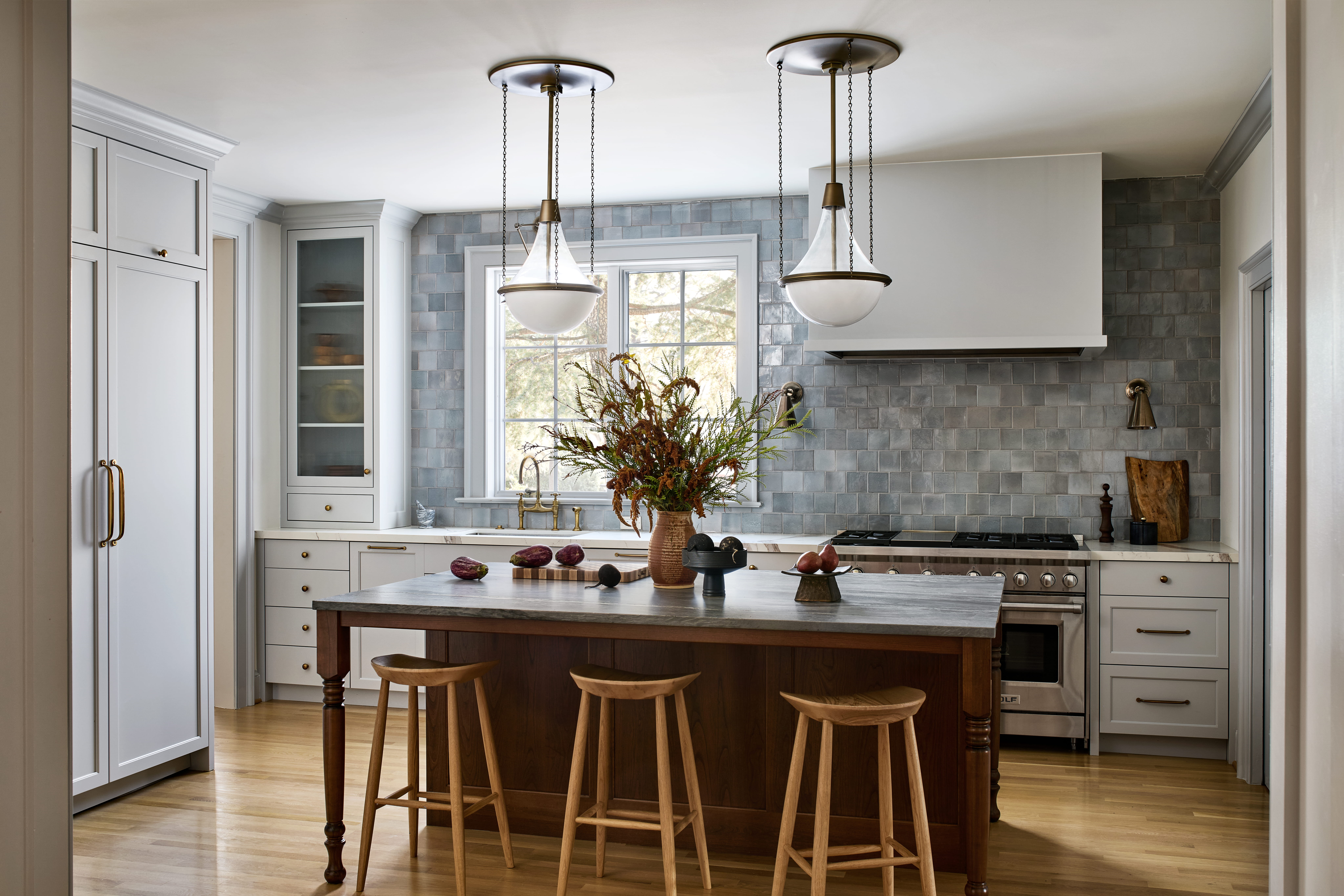 Bright kitchen with white cabinets, a large wooden island, and three wooden stools in a sunlit space with a blue tile backsplash.