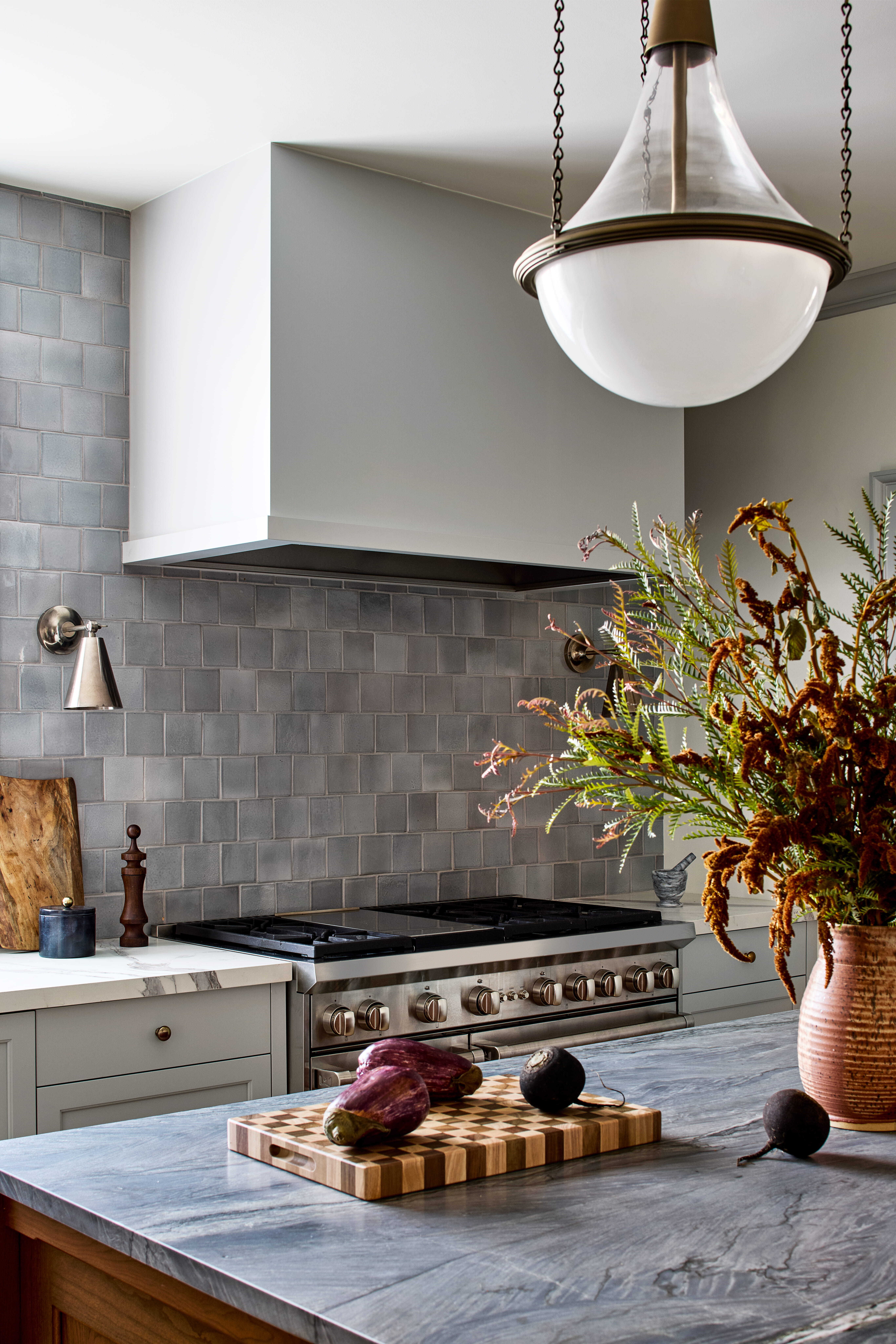 Modern kitchen scene: gray tile backsplash, stainless range, and a marble countertop with a wooden cutting board and eggplants in the foreground.