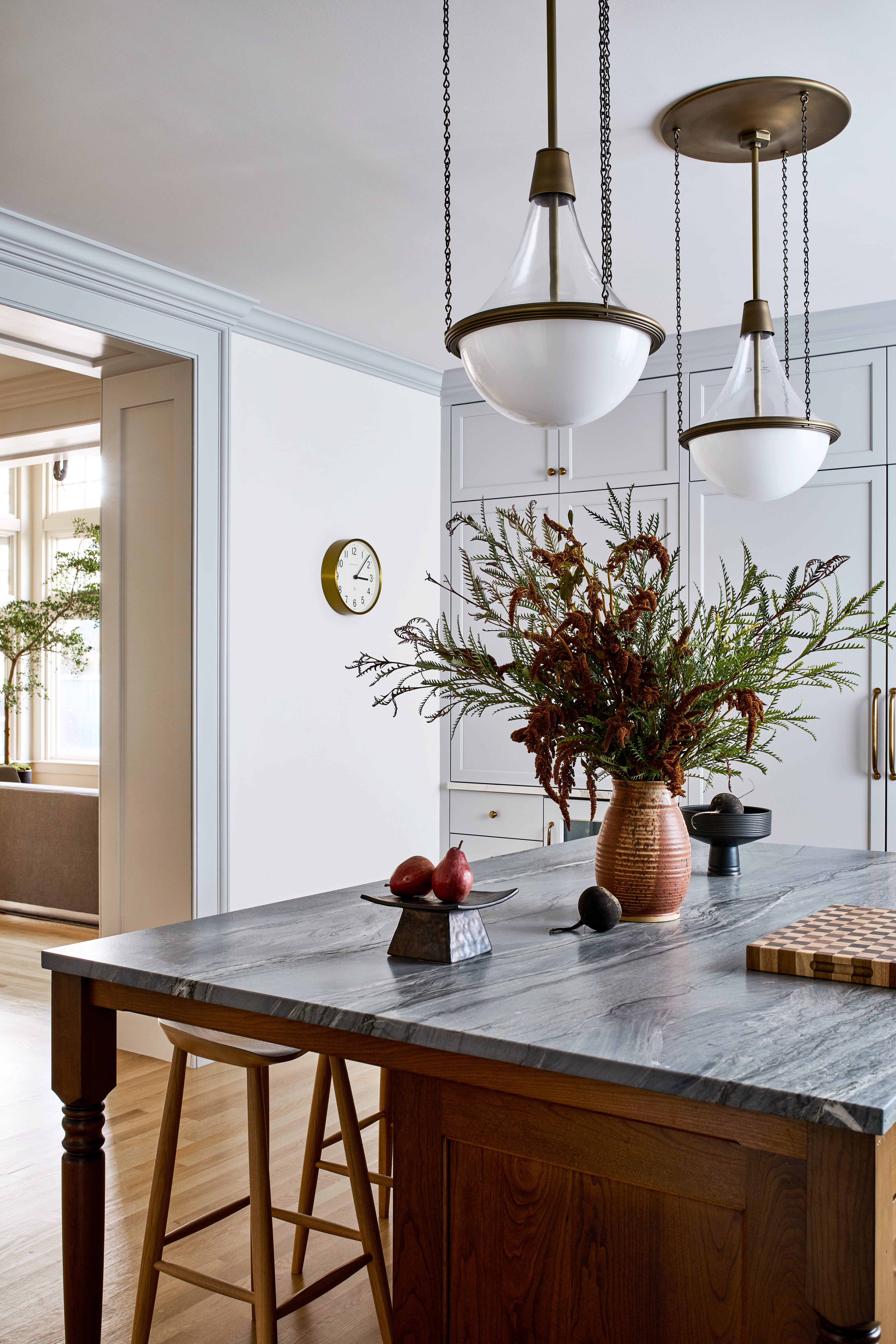 Kitchen island with a dark wooden countertop and wooden stools, a large ceramic vase of dried branches, and fruit on a platter in the foreground.