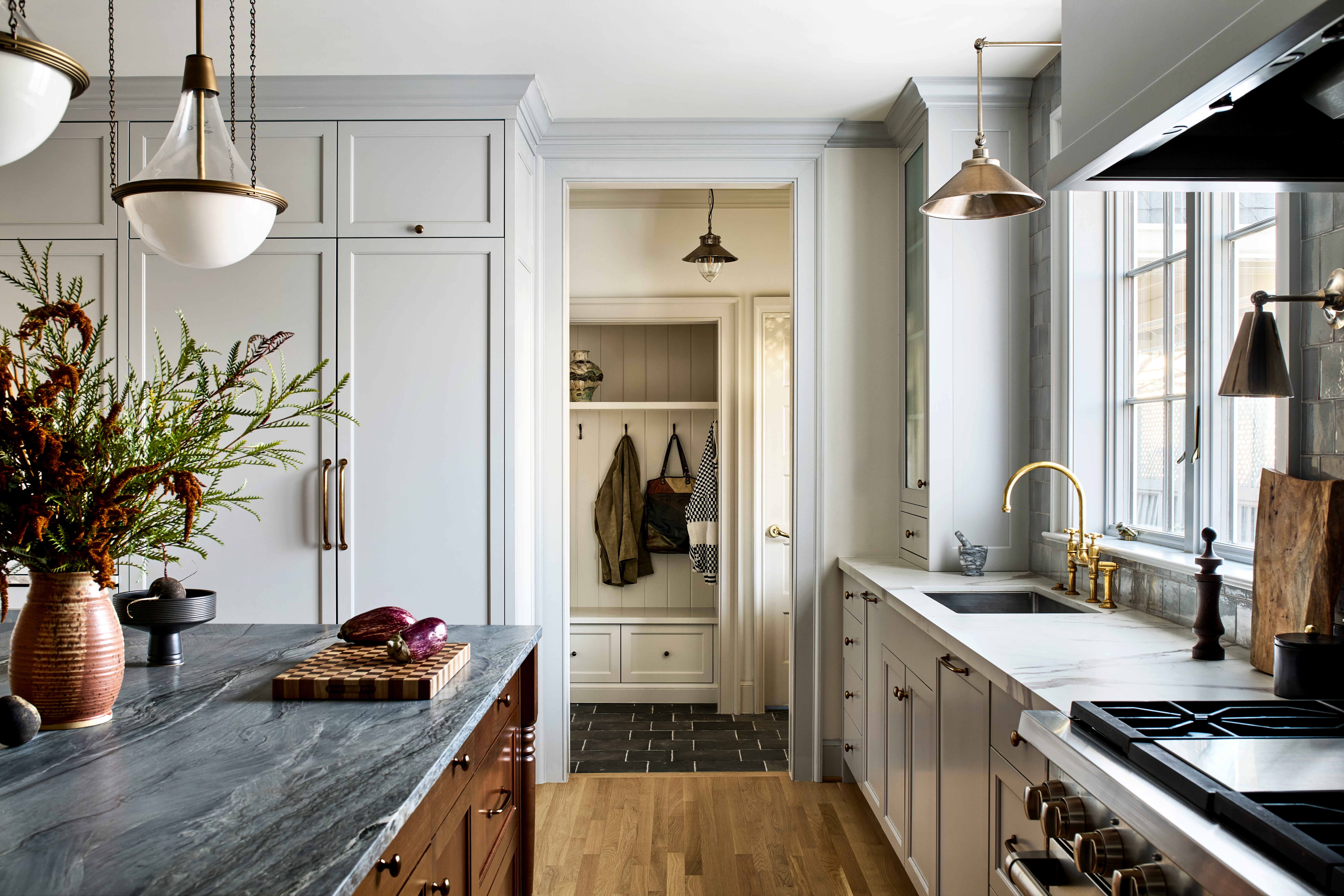 Bright, modern kitchen with gray cabinets, marble countertops, and brass fixtures; leads to a pantry with coats hanging inside.