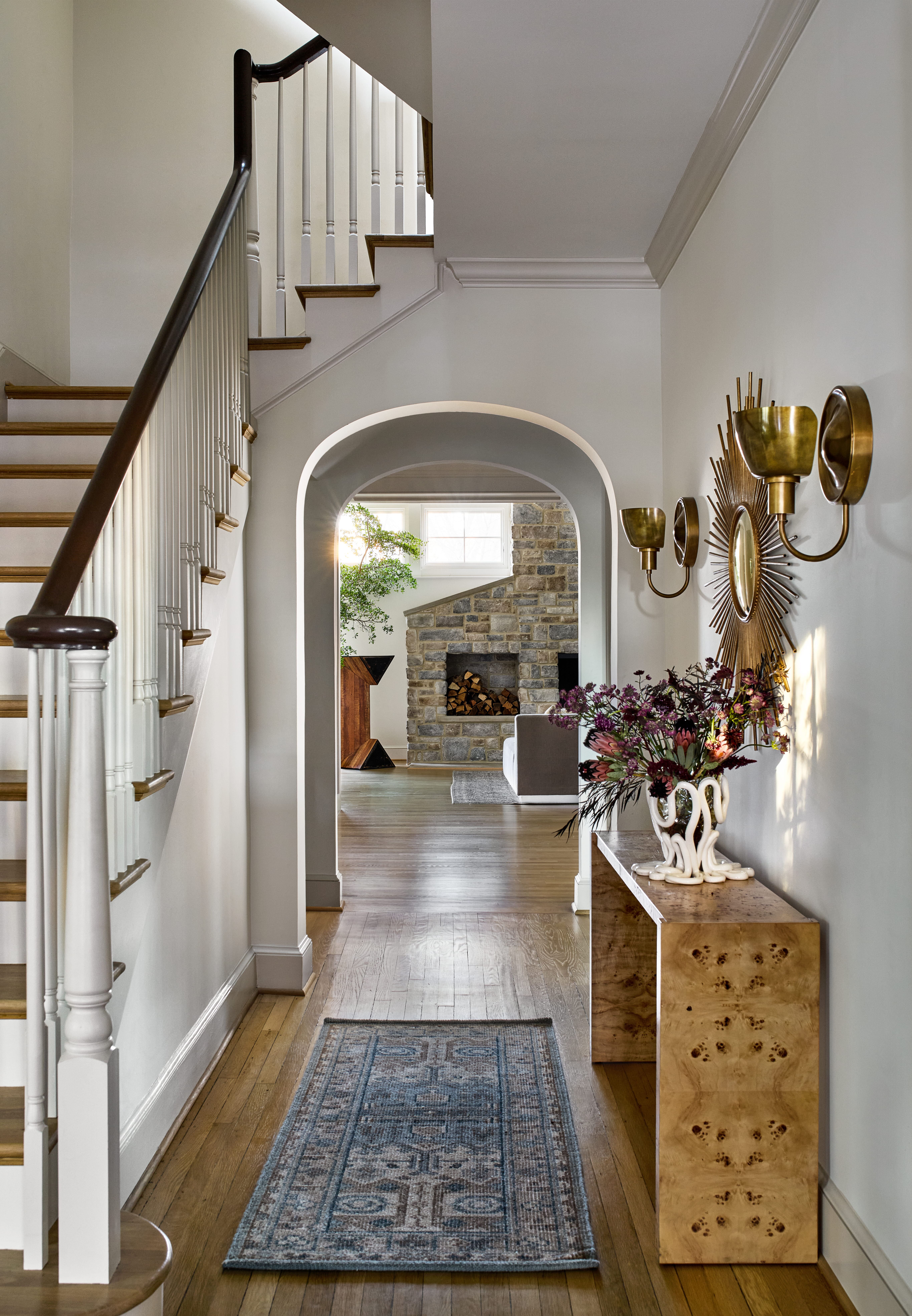 Bright hallway with a curved archway, staircase on the left, and a console table with a vase of purple flowers in the foreground, leading to a living area with a stone fireplace visible through the arch.