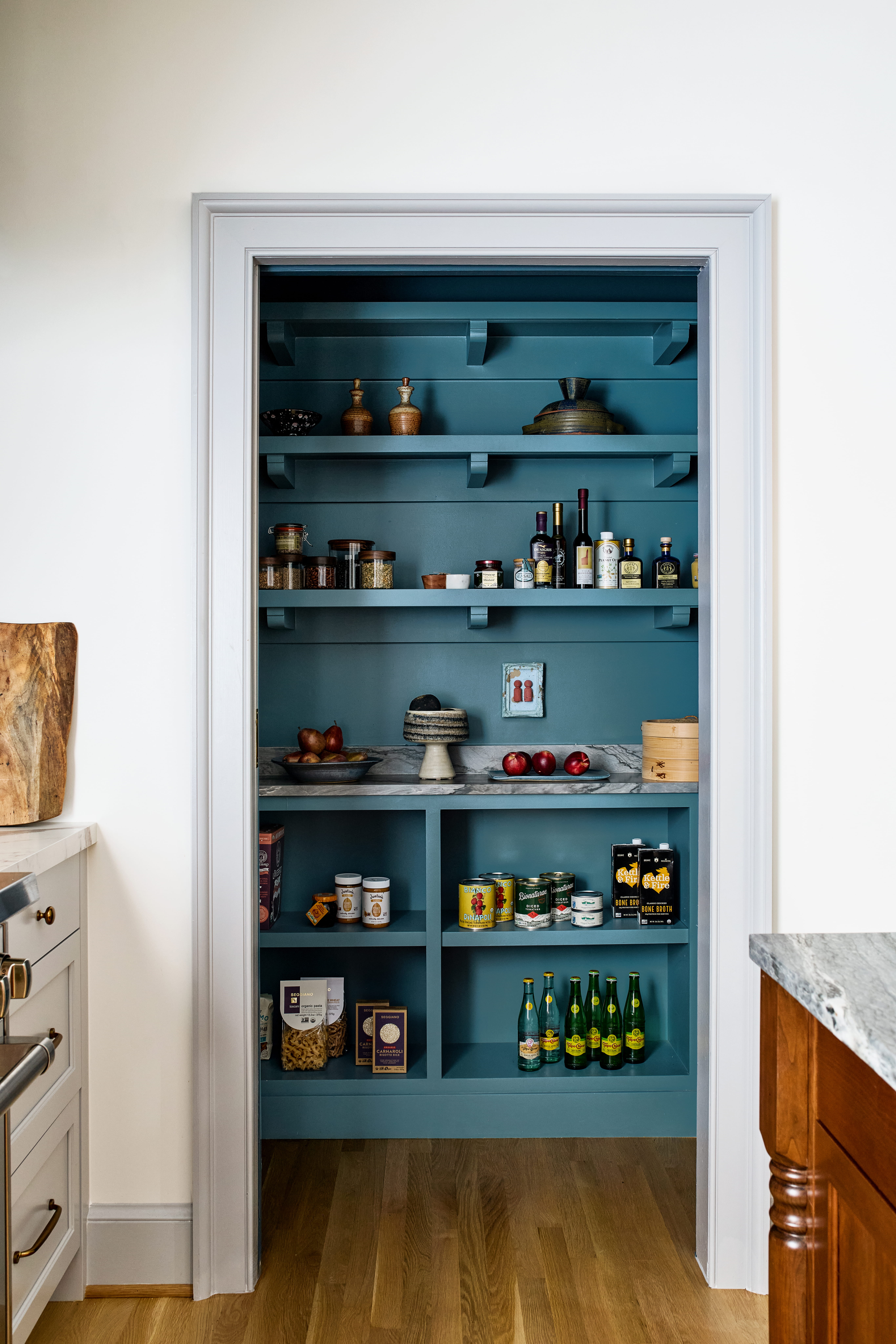 Open pantry shelf painted teal with bottles, jars, and canned goods on blue shelves inside a doorway.