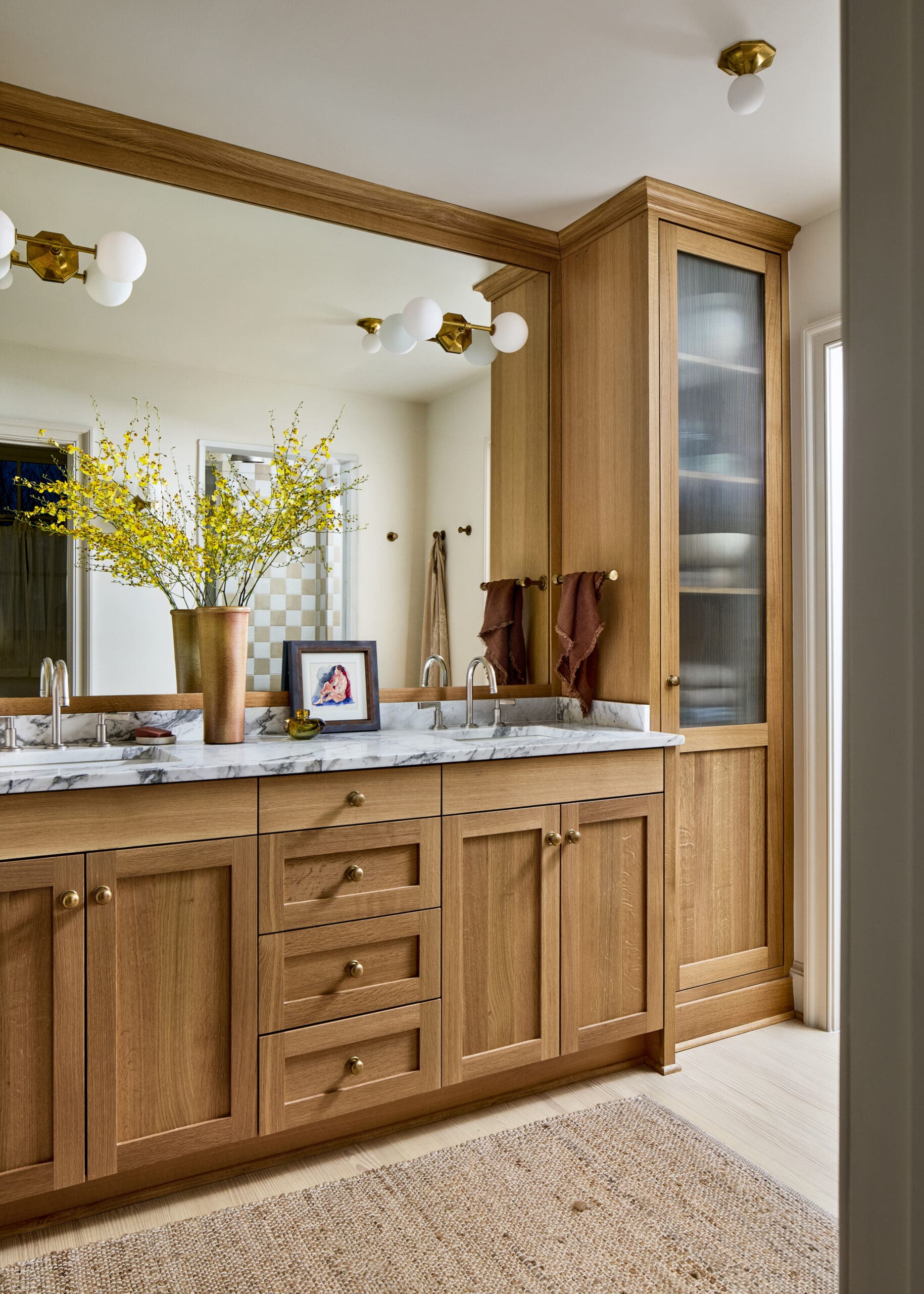 Wooden bathroom vanity with marble countertop and large mirror, yellow flowering branches in a copper vase beside small framed art pieces and beige towels on hooks nearby.