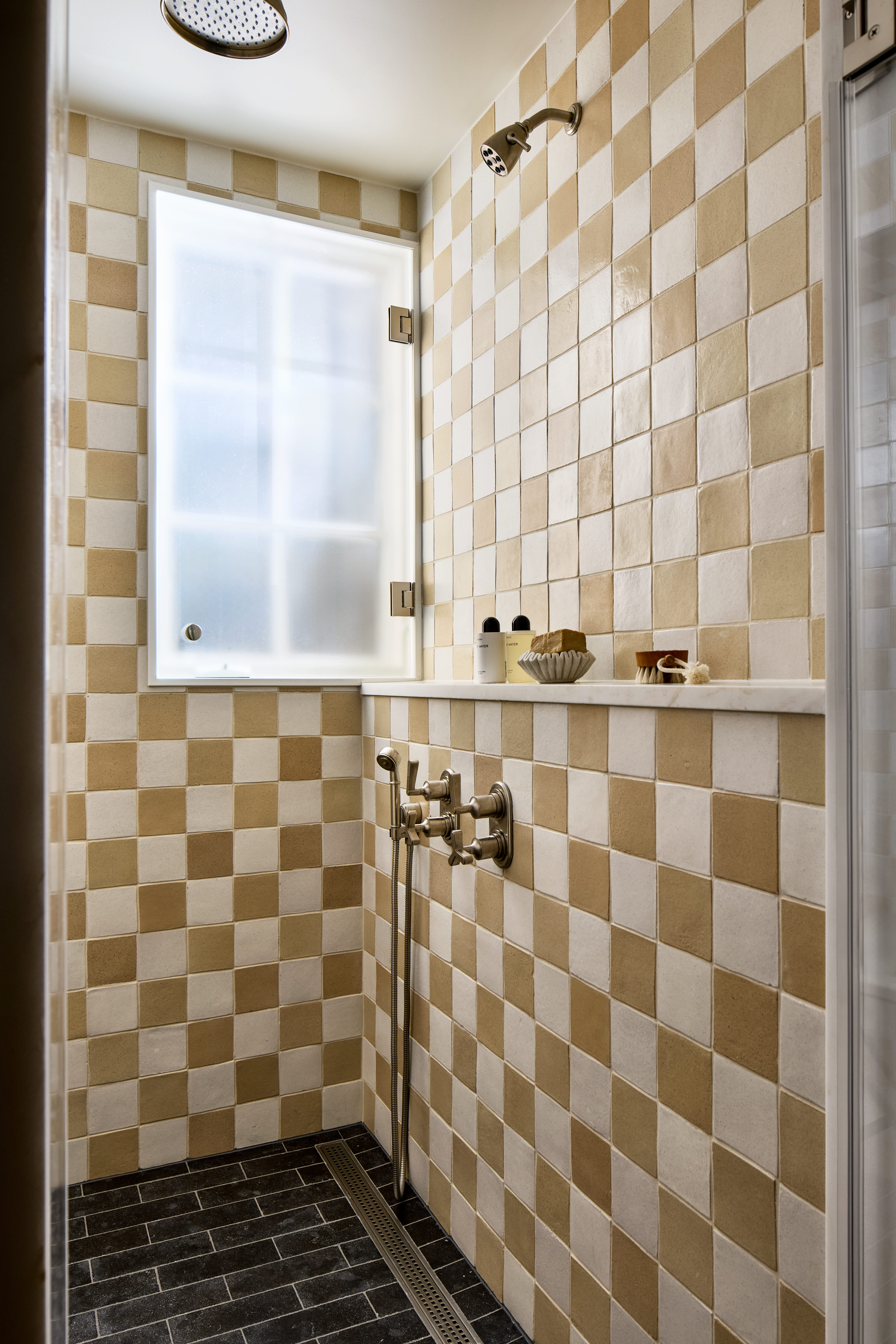 Shower space with beige and cream checkered tile walls, a window, and an overhead rain shower with a handheld head nearby.
