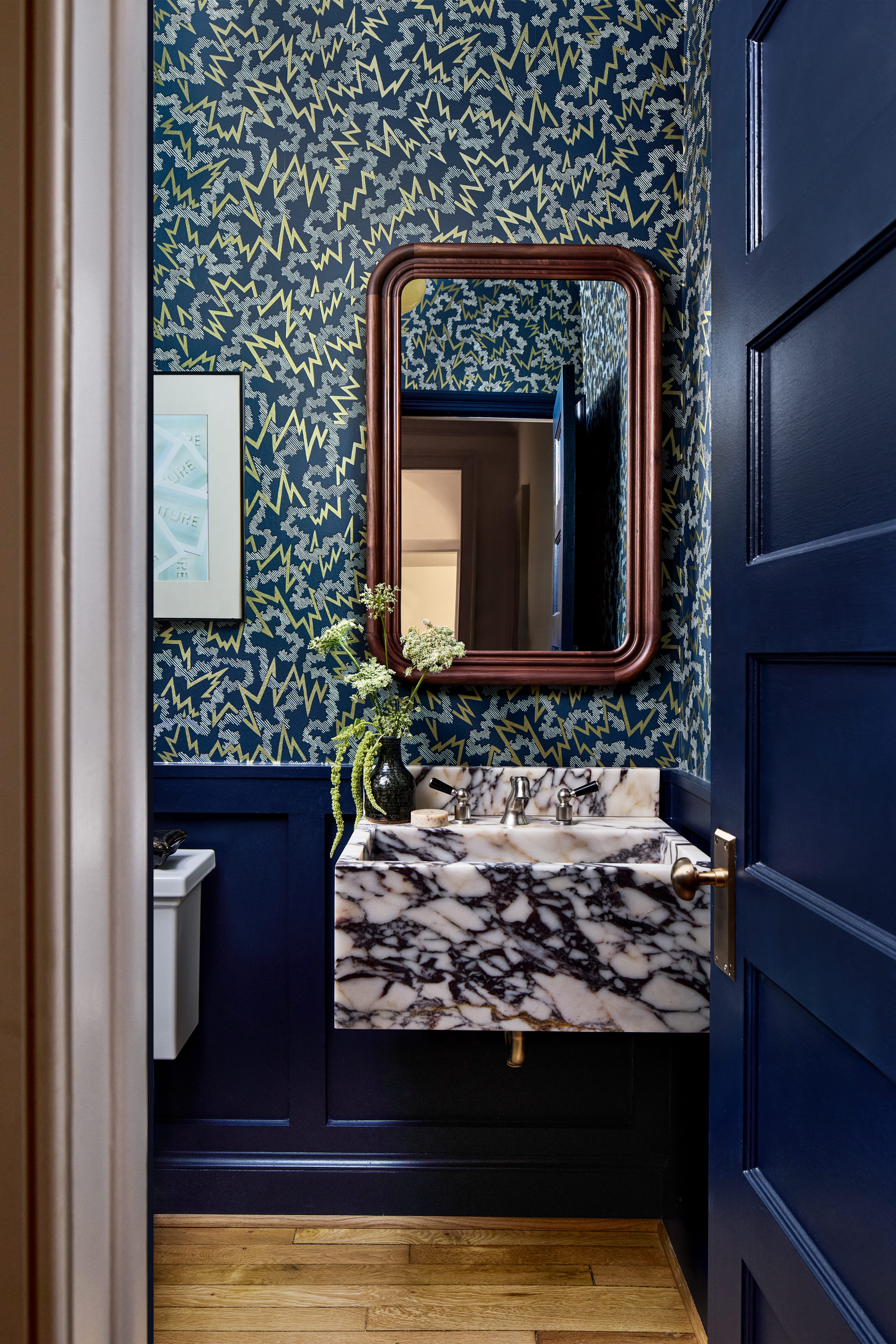 Navy bathroom with zigzag patterned wallpaper, a rounded wooden-framed mirror, and a marble sink with brass fixtures.
