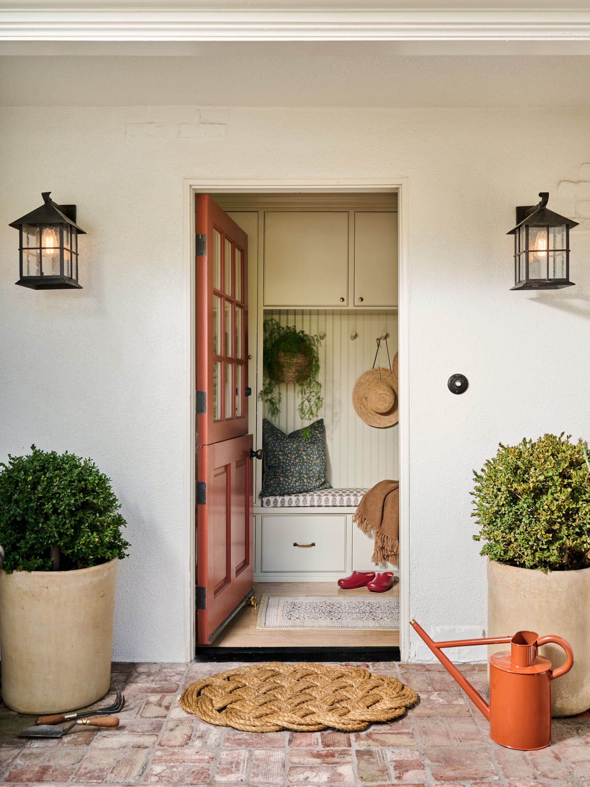 Open front door with red frame leads into a cozy mudroom: built-in cabinets, bench with cushions, hanging straw hat, and green plant.