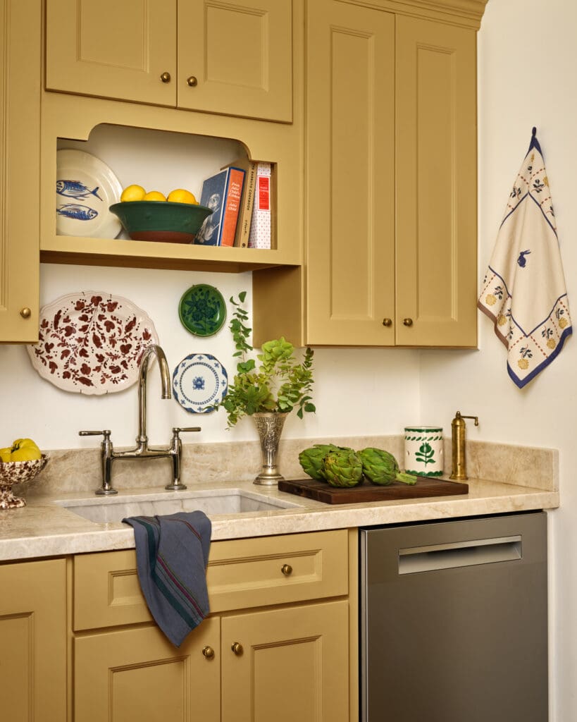 Beige kitchen cabinets with an open shelf holding a green bowl of lemons, ceramic plates, and cookbooks beside the sink area.